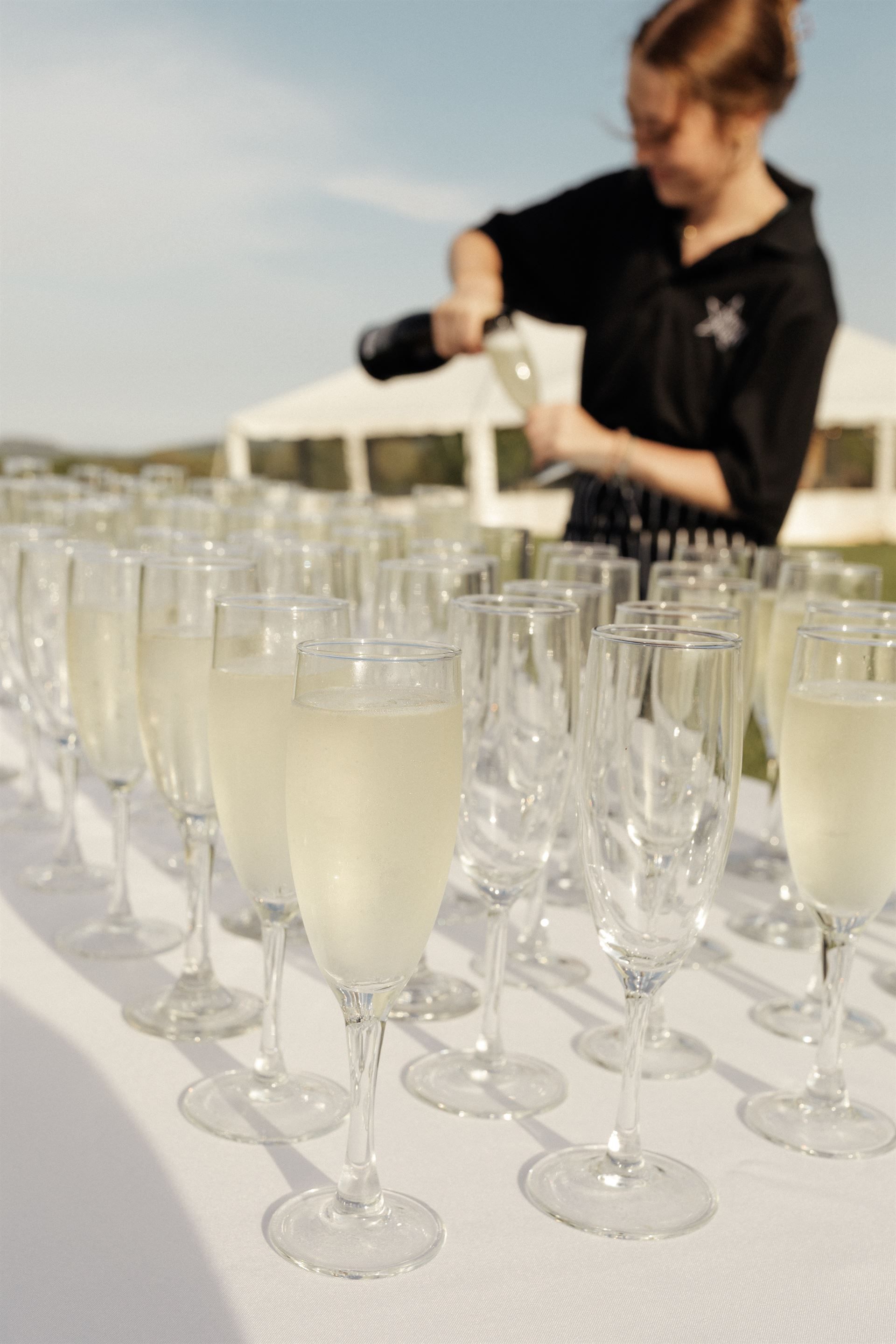 A person pours champagne into fluted glasses on a white table outdoors, tent in the background.