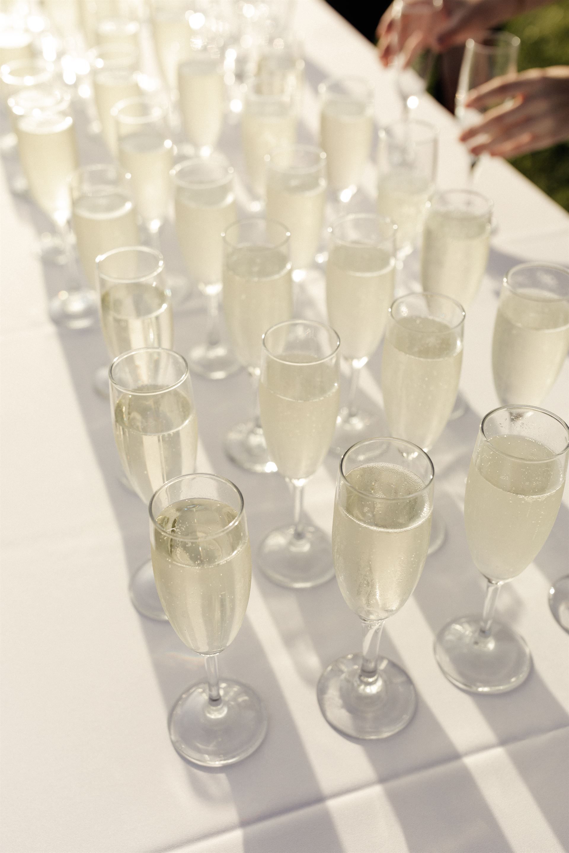 Champagne flutes arranged on a white tablecloth, being served; two hands reach for a glass.