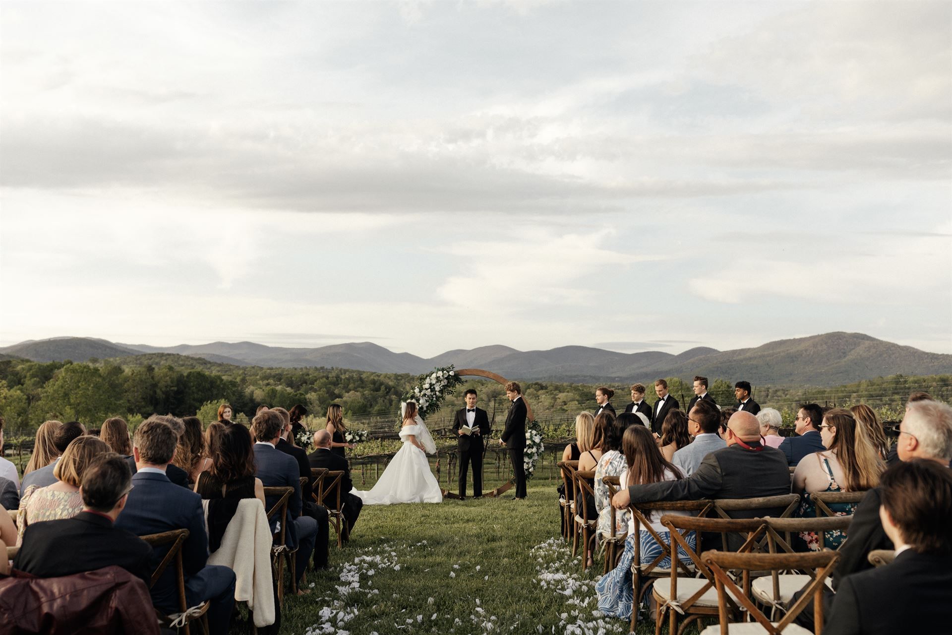 Wedding ceremony outdoors, bride and groom exchanging vows, guests seated, scenic mountain backdrop.