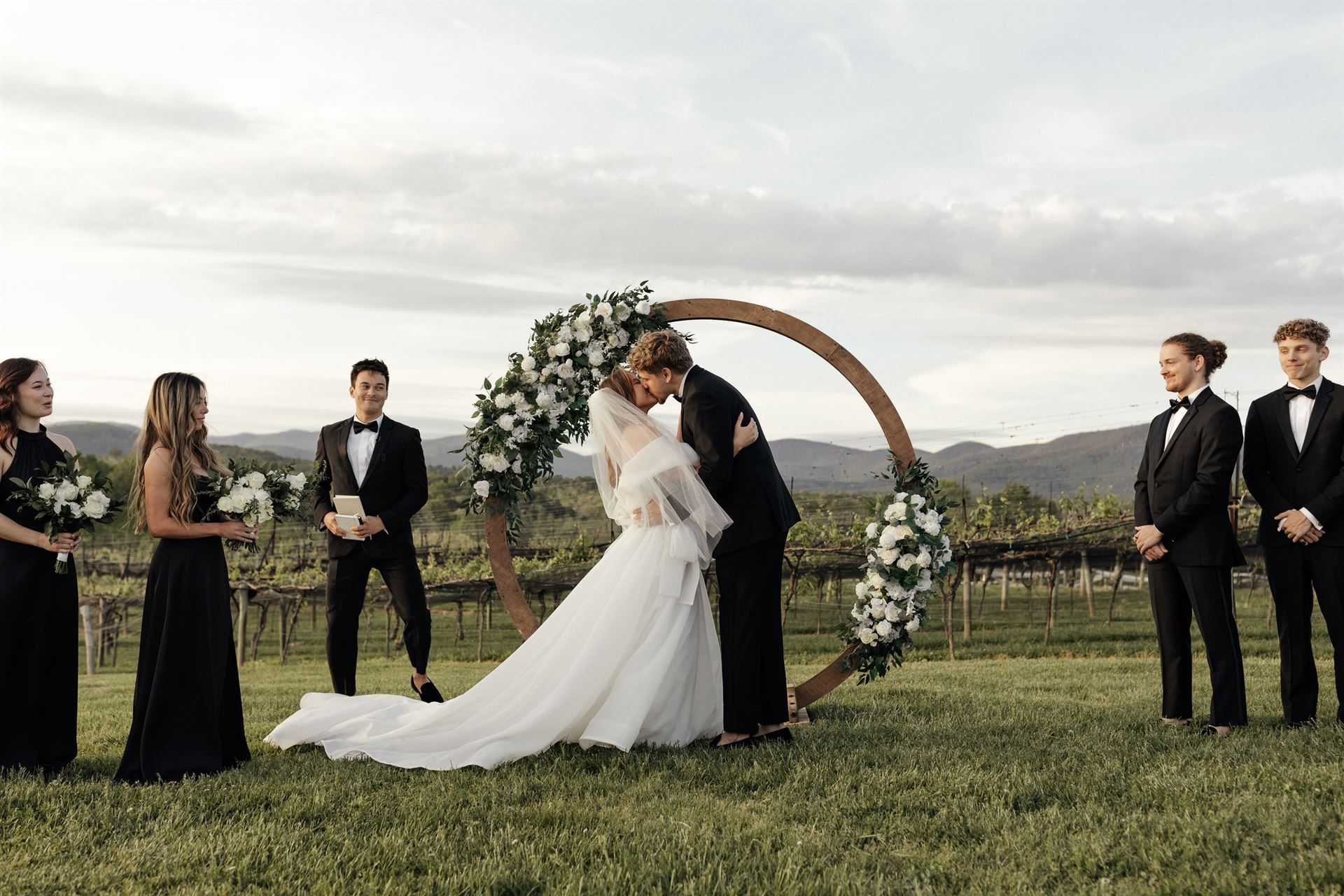 Wedding ceremony with couple kissing under floral arch, surrounded by wedding party in a vineyard.