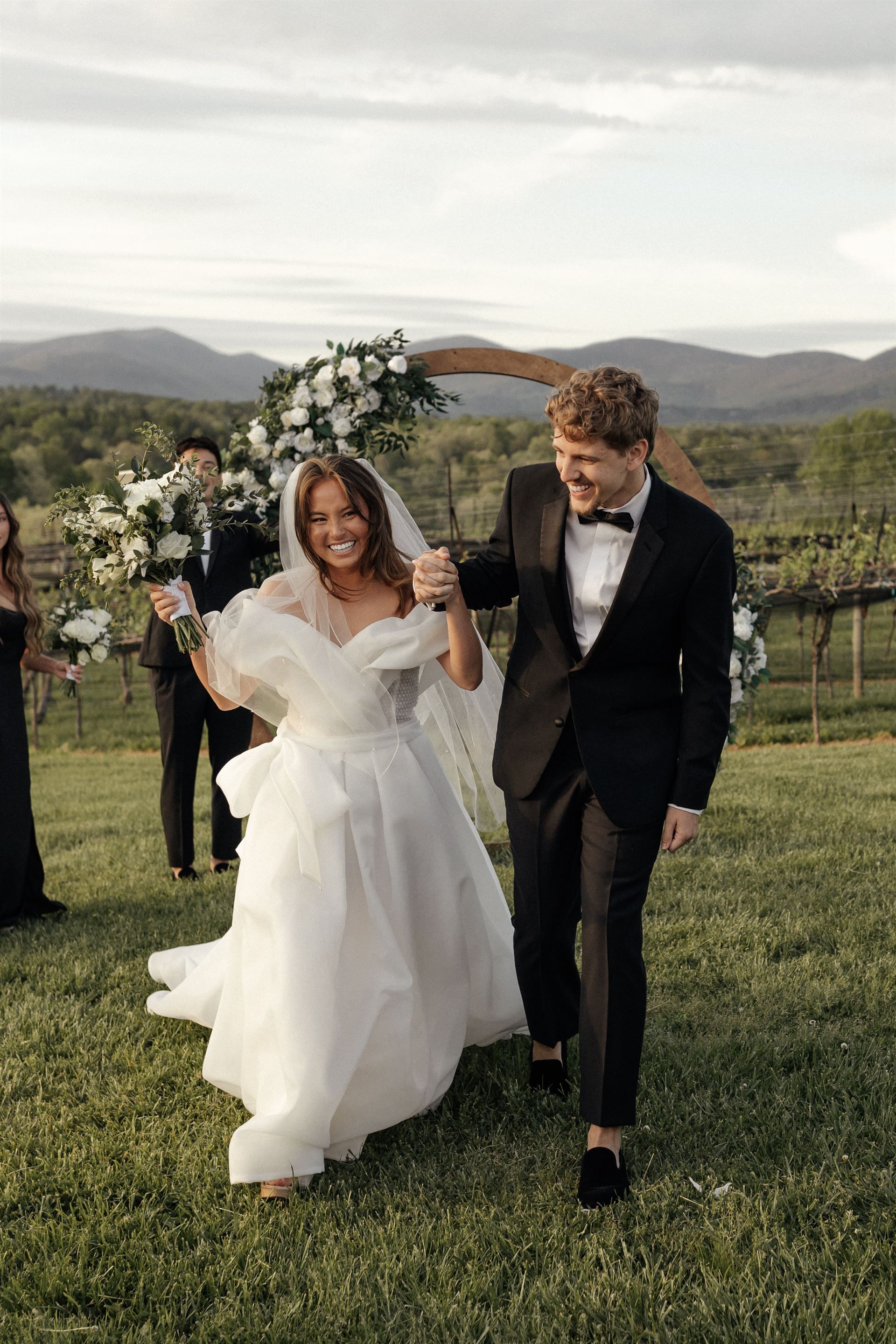 Newlyweds walk away from ceremony, holding hands, with a flower arch and vineyard backdrop.