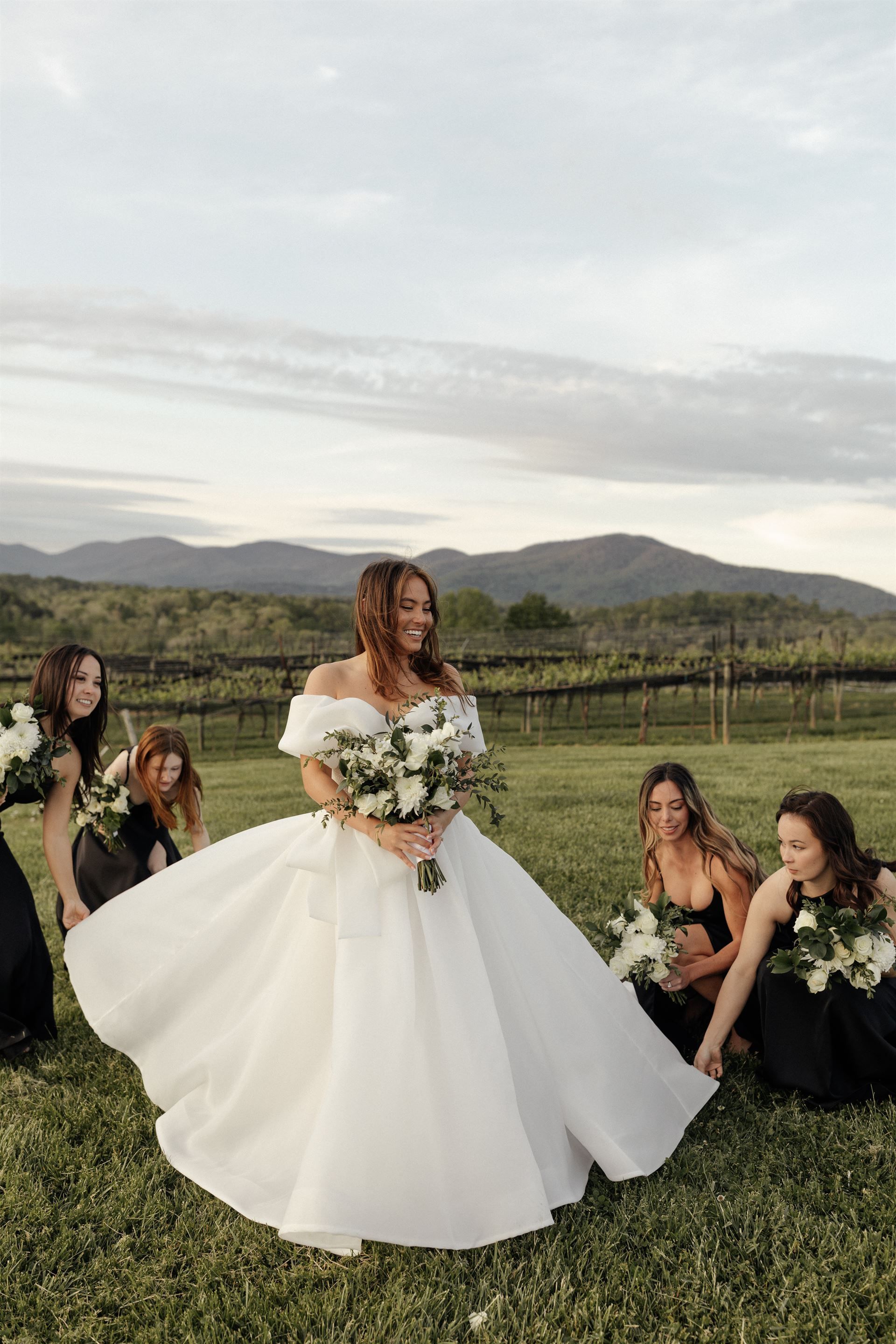 Bride in white gown surrounded by bridesmaids in black, holding bouquets, in a field with mountains.