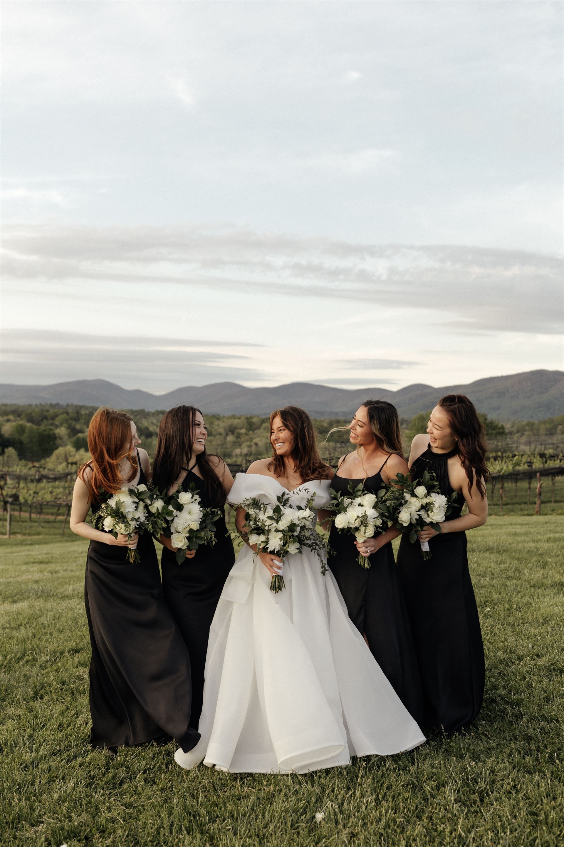 Bride with four bridesmaids holding bouquets, all smiling on a grassy hill with mountains in the background.