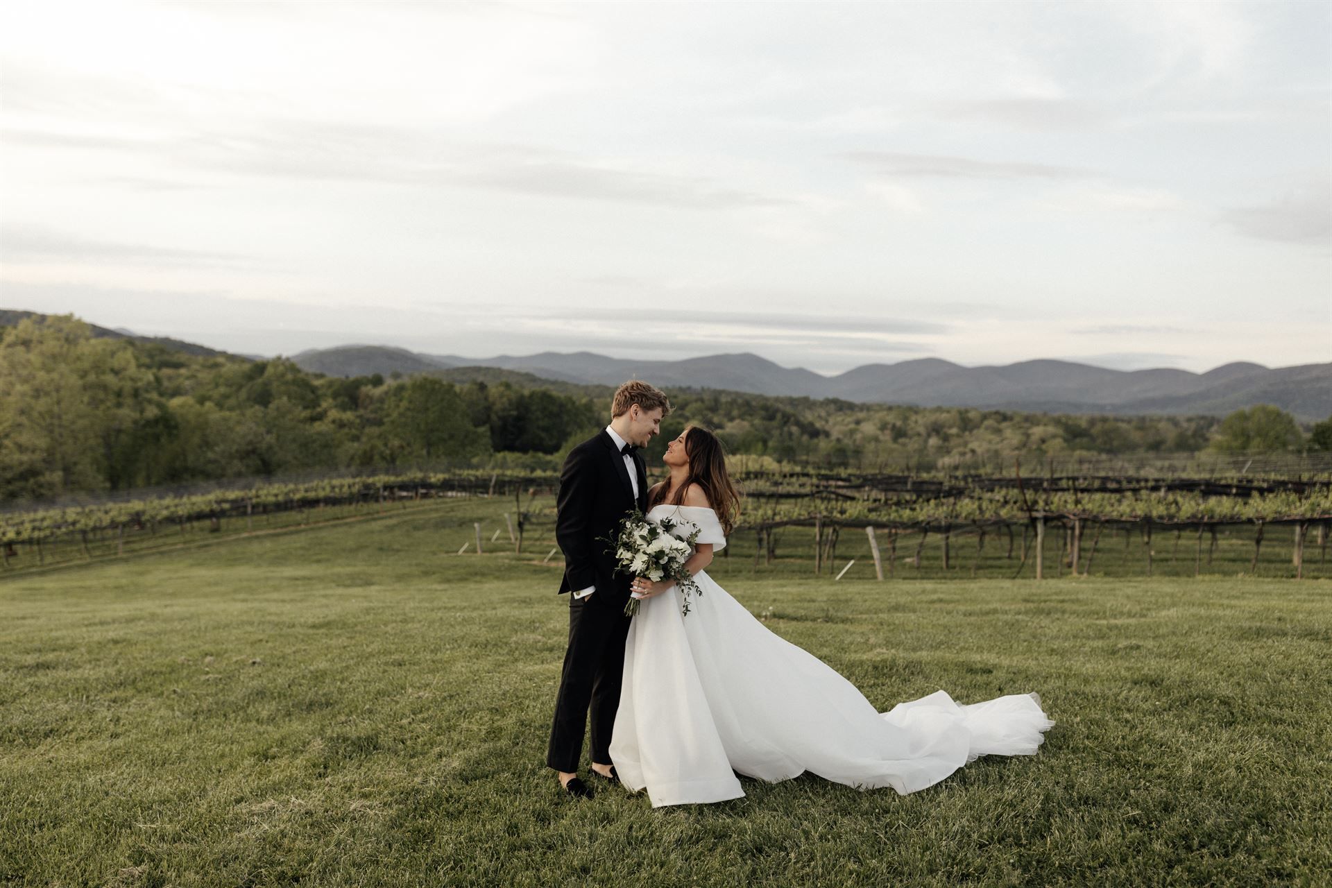 Couple embracing in a field with a vineyard and mountain backdrop. Bride in white dress, groom in black tux.