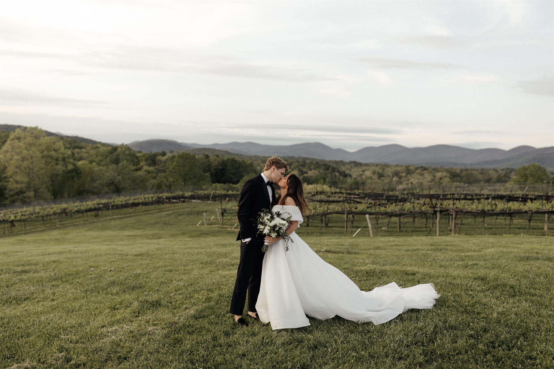 Couple kissing outdoors, bride in white dress, groom in black suit, mountain backdrop.