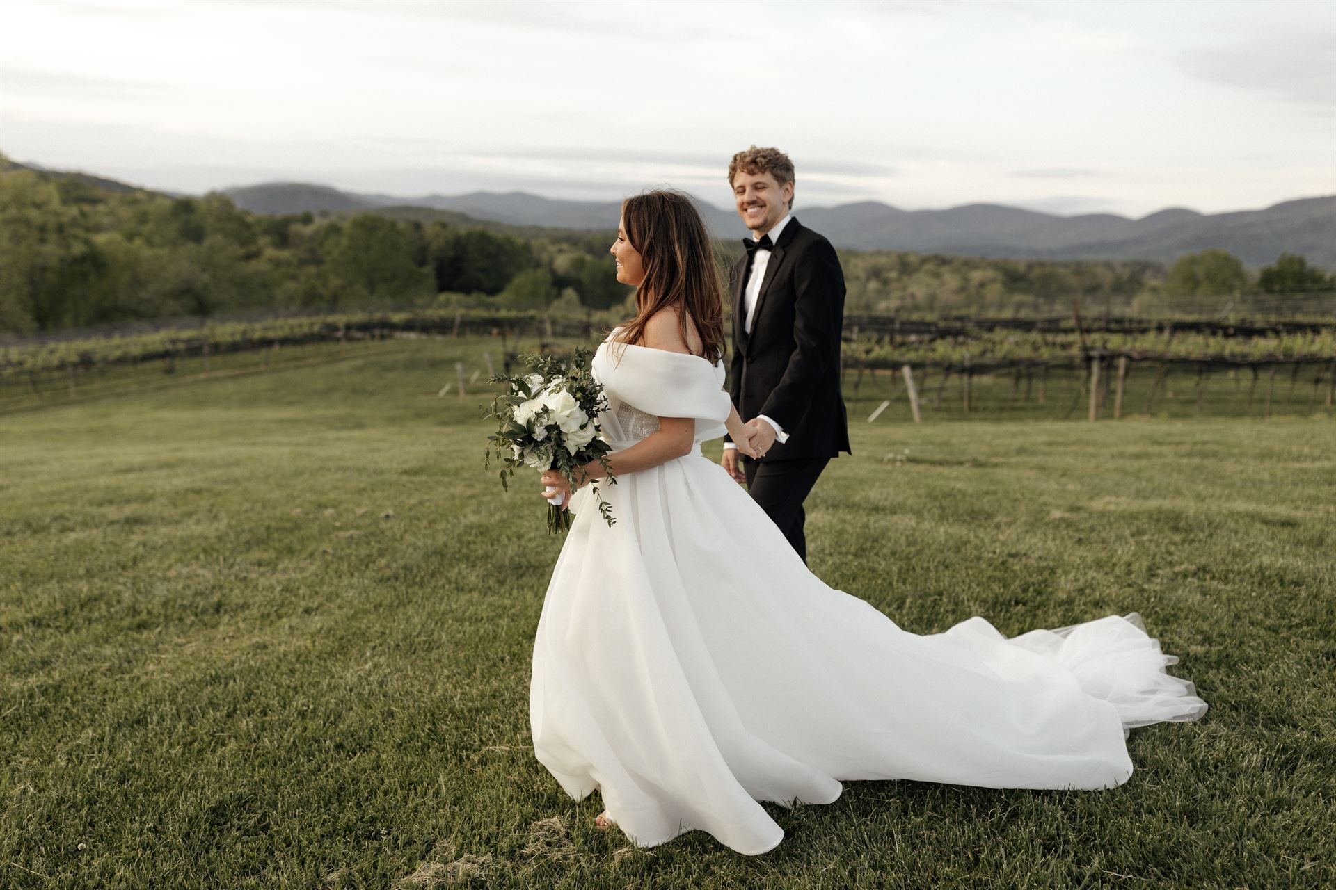 Bride and groom walk in a vineyard, mountains in the background. Bride in white gown with bouquet. Groom in black tuxedo.