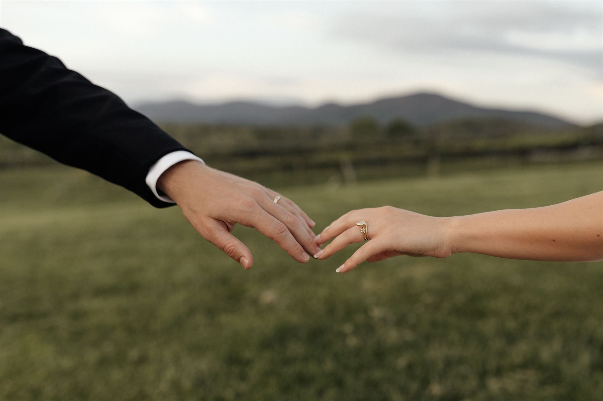 Hands of a couple touching, wearing wedding rings, with a field and mountains in the background.