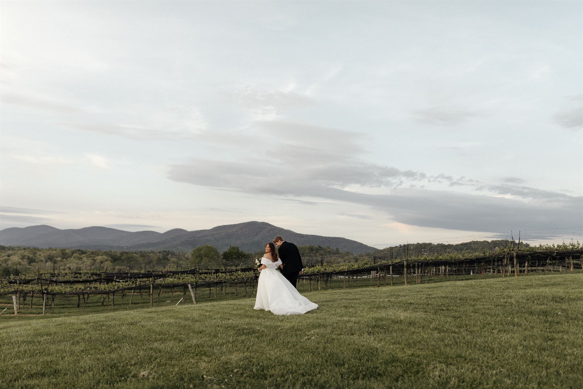 Bride and groom embrace on a grassy hill overlooking a vineyard and mountains at sunset.