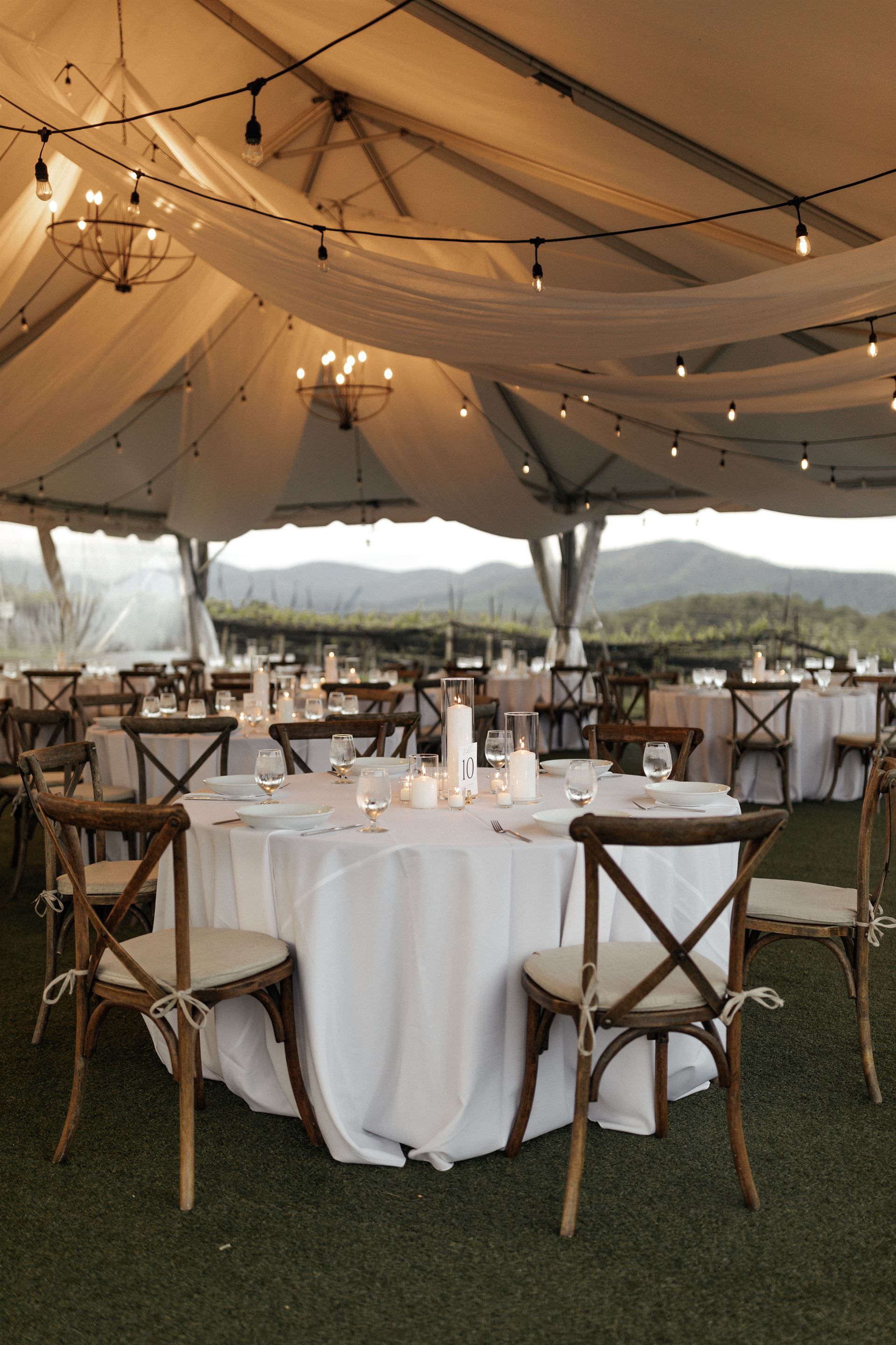 Wedding reception setup under a tent with tables, chairs, and string lights, with a mountain backdrop.