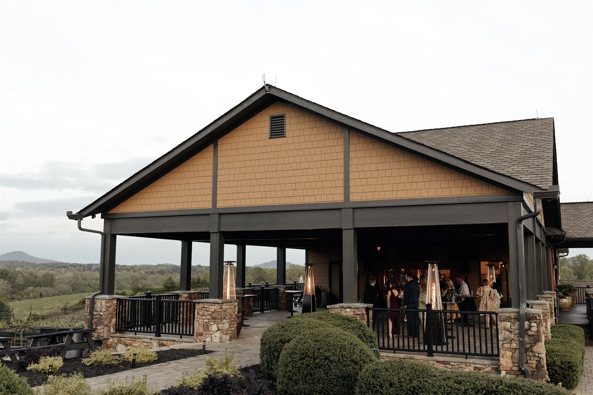 Covered outdoor patio with people, overlooking a landscape. Brown building with dark trim.