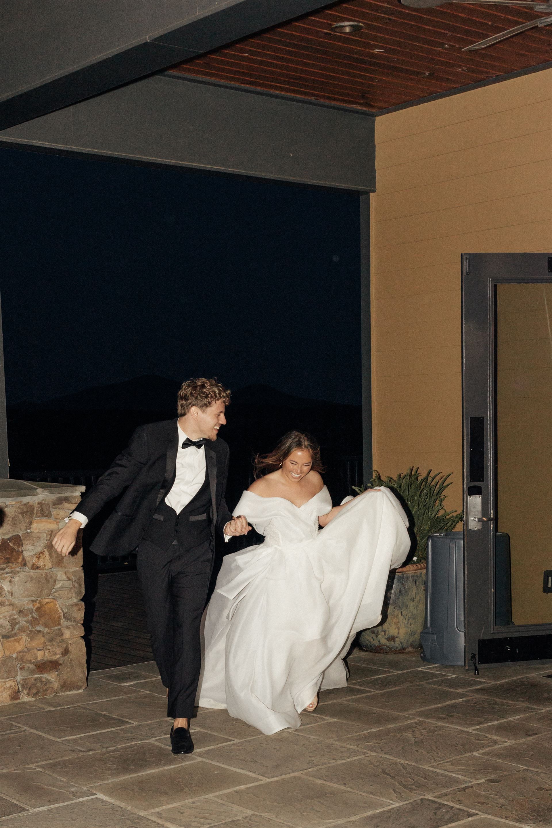 Newlyweds run outside at night, groom in tuxedo, bride in white gown, holding hands.