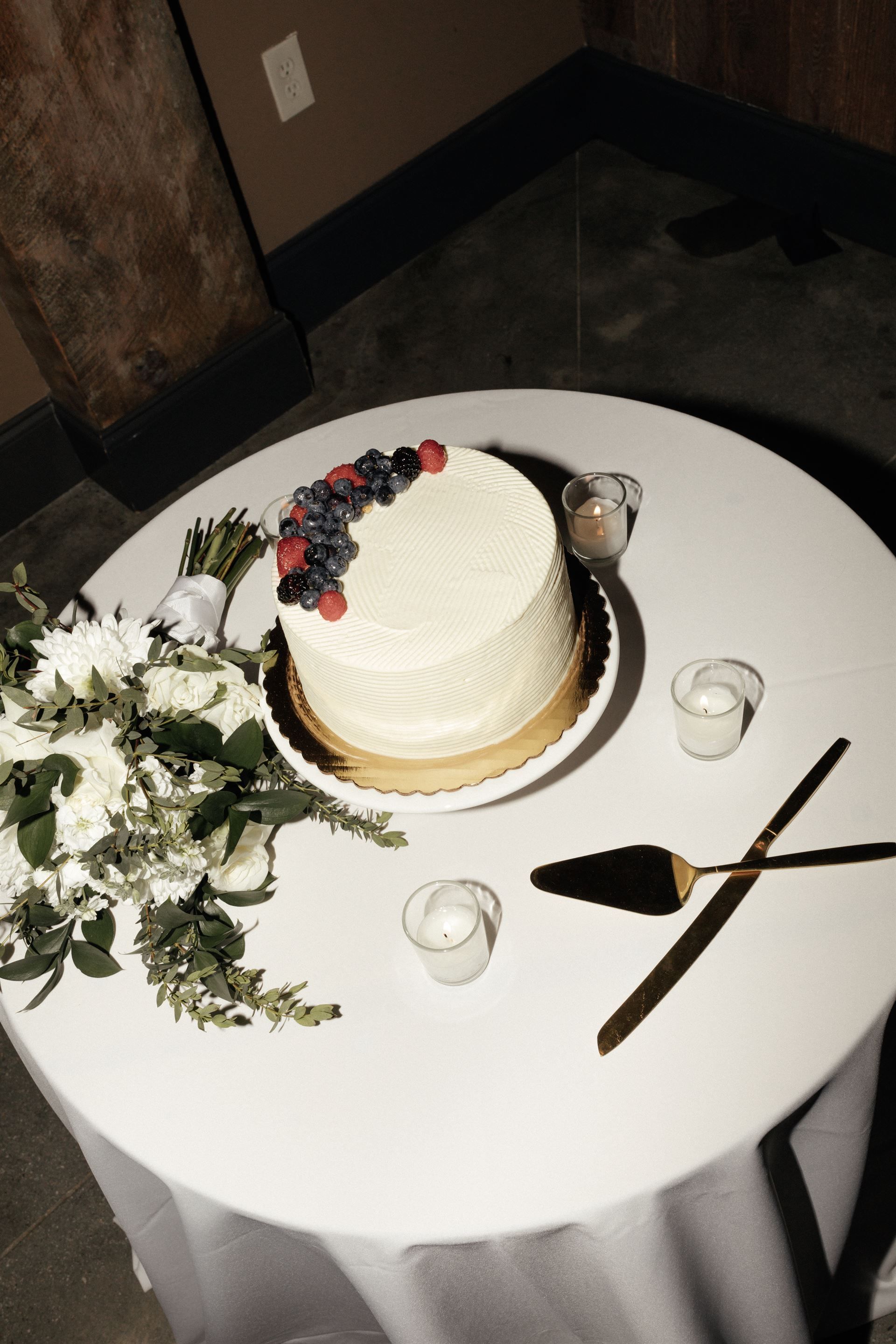 Wedding cake decorated with berries on a round table, next to flowers and candles.