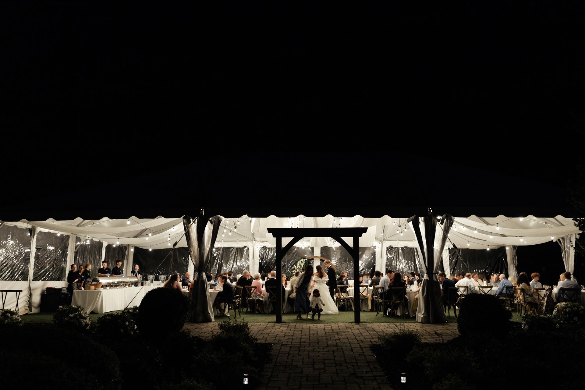 Wedding reception at night under clear tents; bride and groom centered under an archway, guests seated at tables.