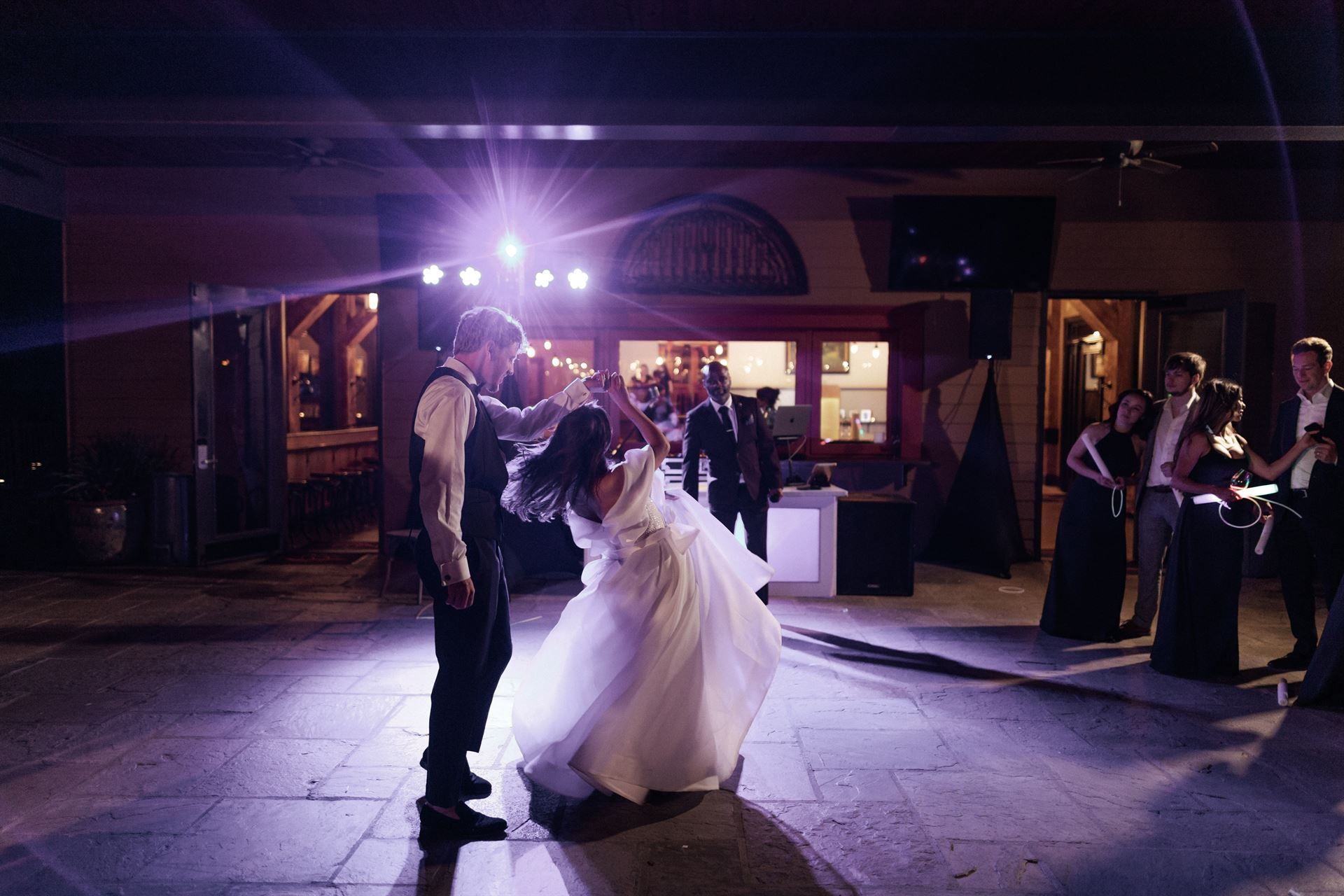 Couple dancing at wedding reception. Bride in white dress and groom in vest. Purple spotlights illuminate dance floor.