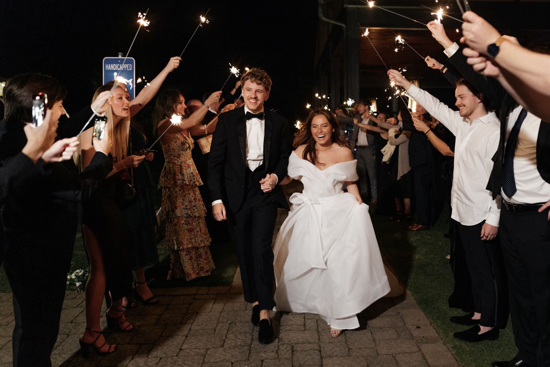 Newlyweds run through a sparkler exit at night, guests cheering, with a building backdrop.