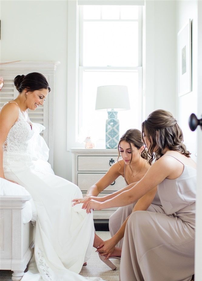 Bride being helped into her dress by two attendants in a bedroom, near a window.