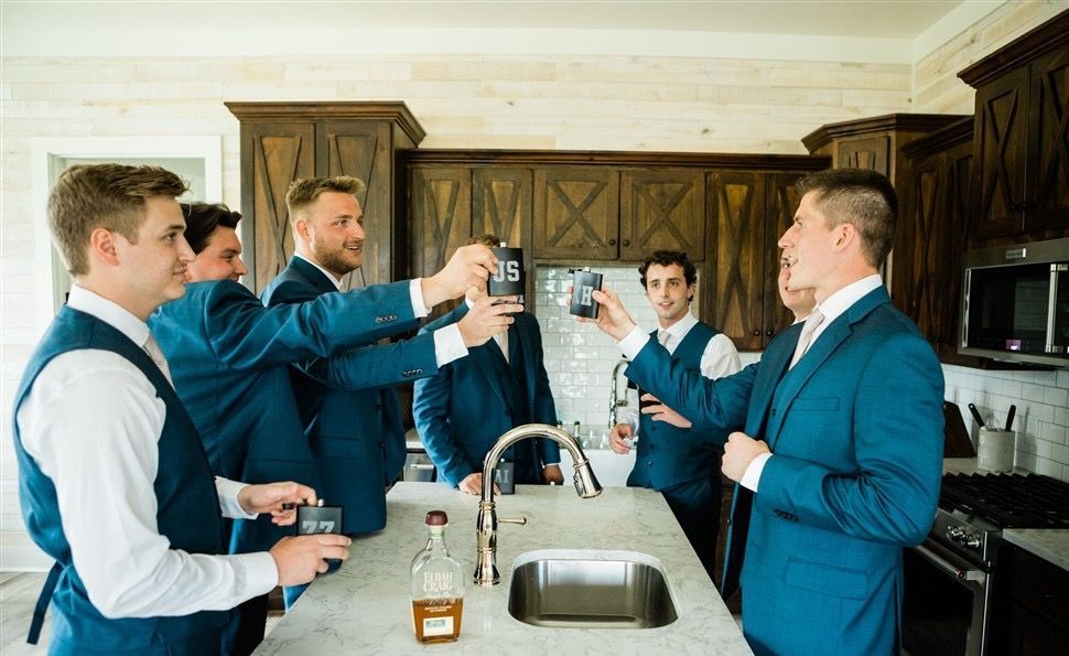 Men in suits toasting with drinks in a kitchen, celebrating.