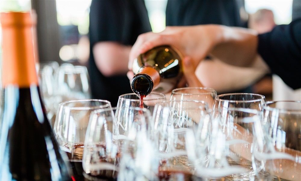 Wine being poured into multiple glasses at a tasting. Red liquid, close-up shot, in a brightly lit setting.