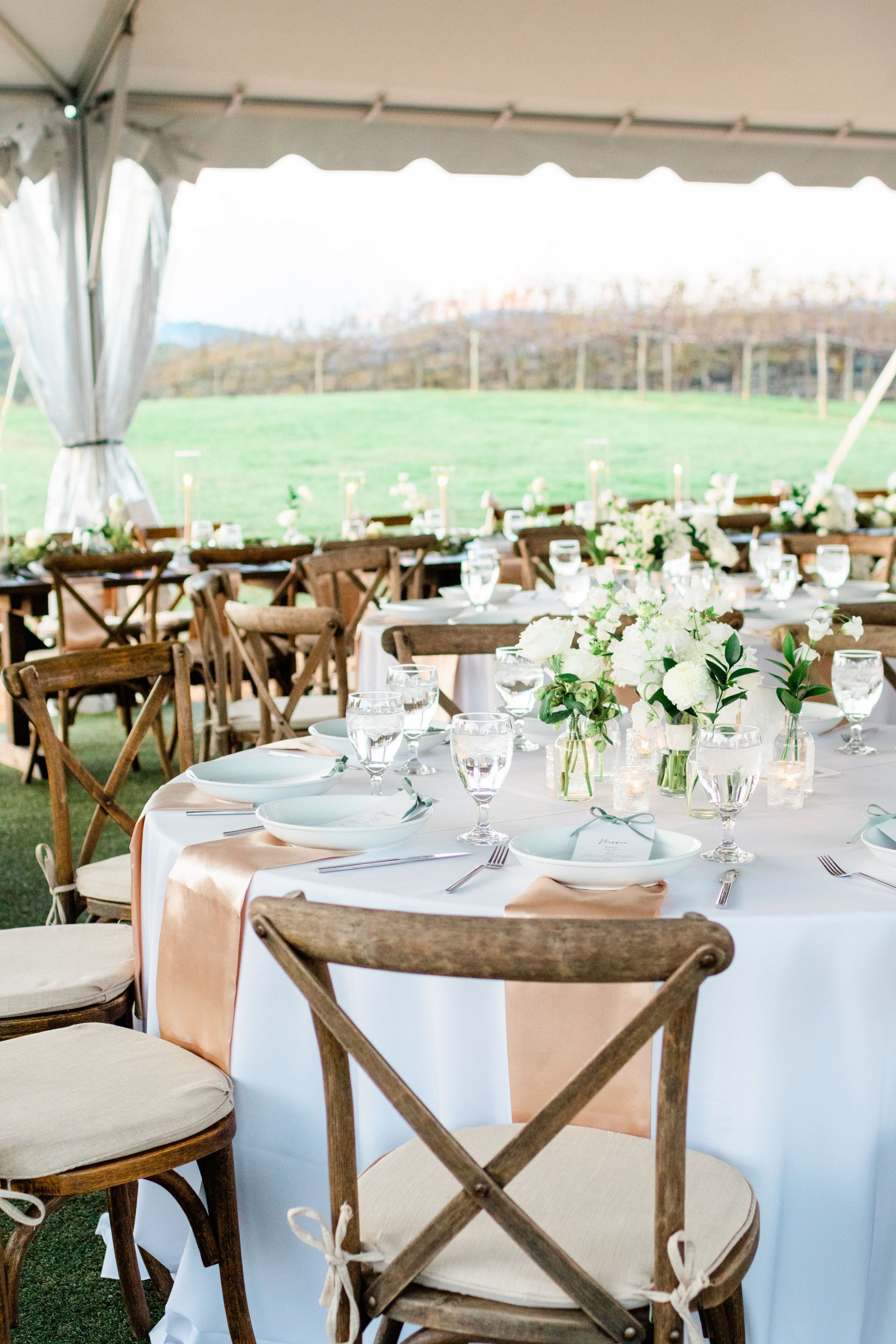 Wedding reception table setting under a white tent; round table with white tablecloth, flower centerpiece, peach runner, wooden chairs.