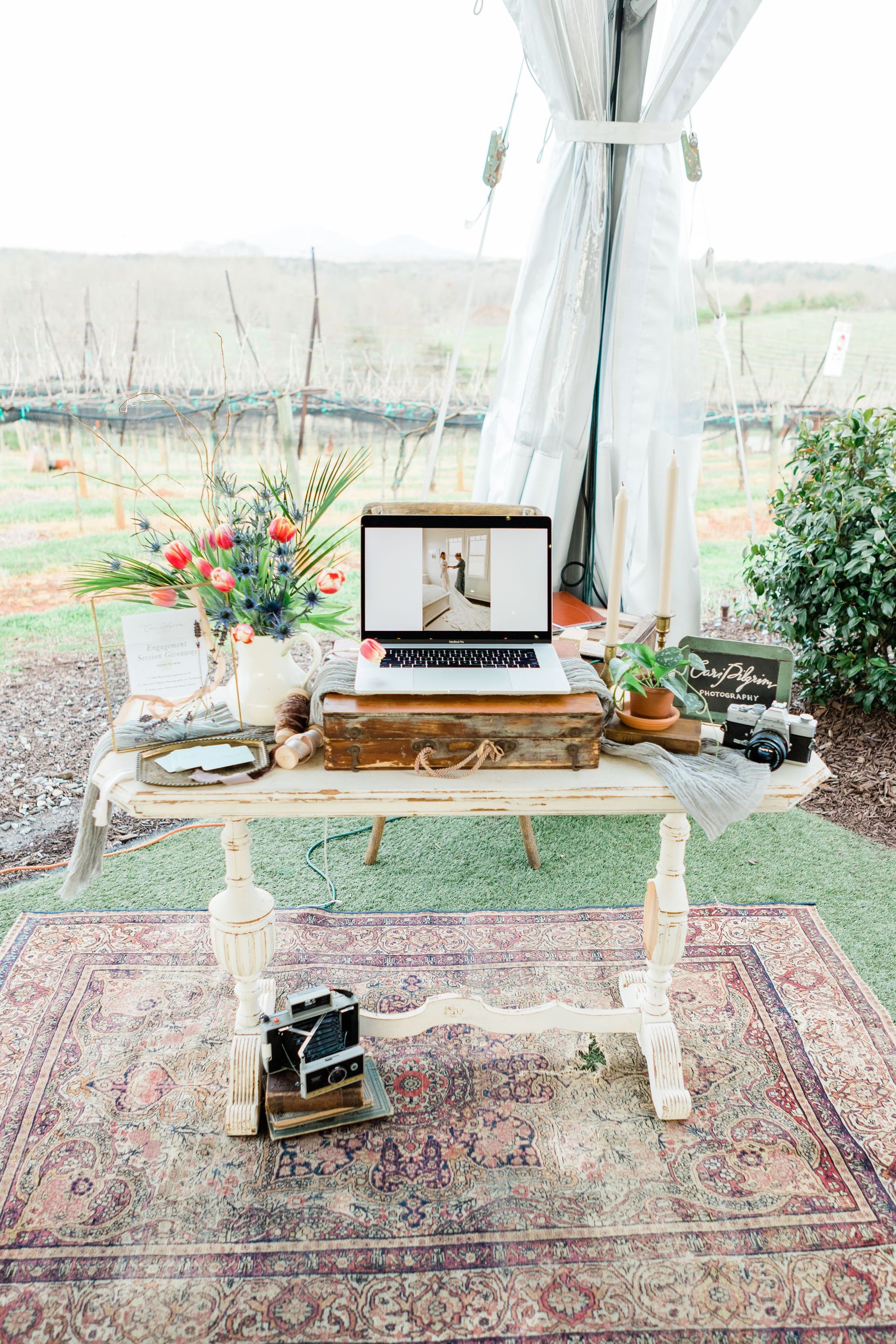 A laptop on a rustic table with decor, outdoors in a tent setting. Cameras, rug, and flowers complete the scene.