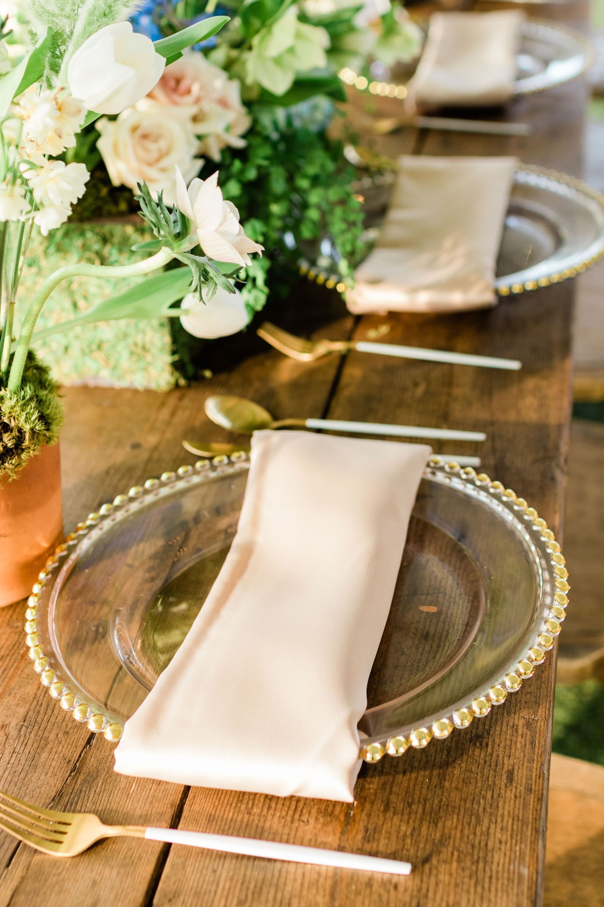 Table setting with gold-rimmed charger plate, gold flatware, and folded beige napkin on a wooden table. Floral arrangement.