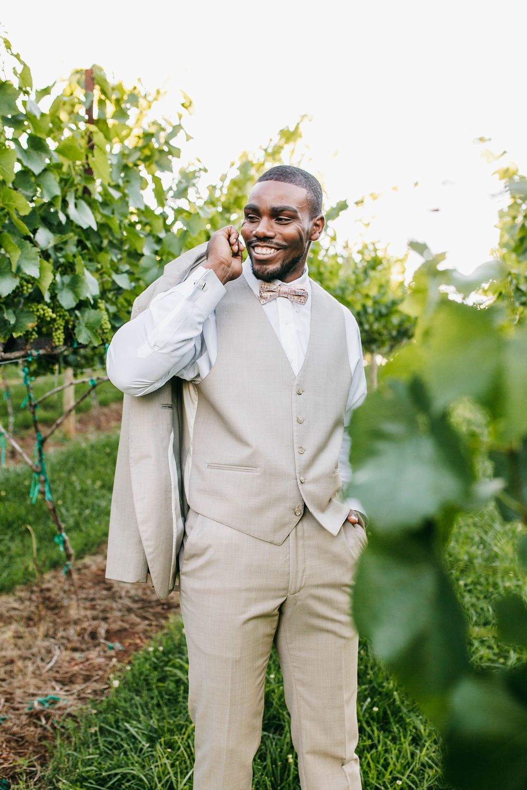 Man in a tan suit and bow tie smiles, holding his jacket and standing in a vineyard.