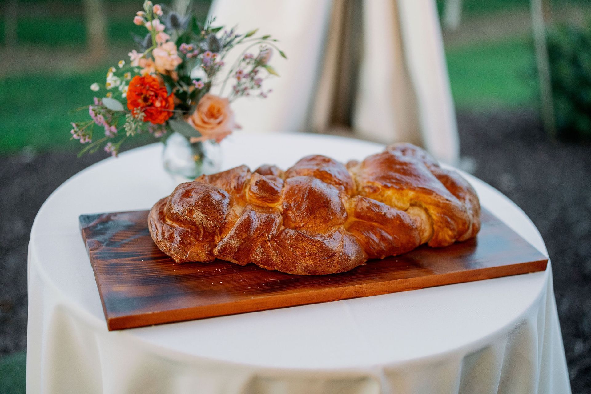 Challah bread loaf on a wooden board atop a white round table, with flowers in the background.