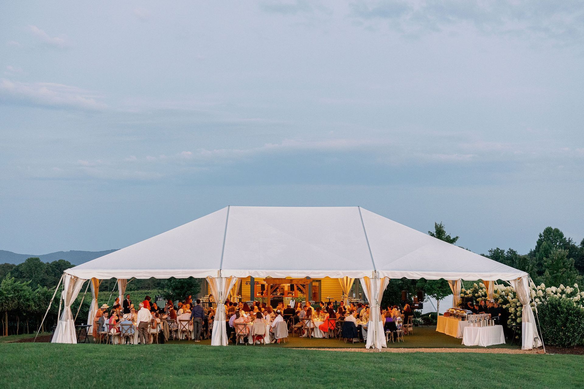 White tented outdoor wedding reception with guests seated at tables, a backdrop of mountains.