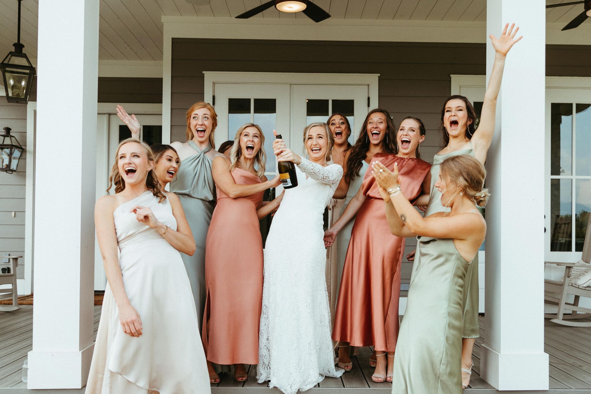 Bride and bridesmaids cheering on a porch; bride pops champagne, excited expressions.