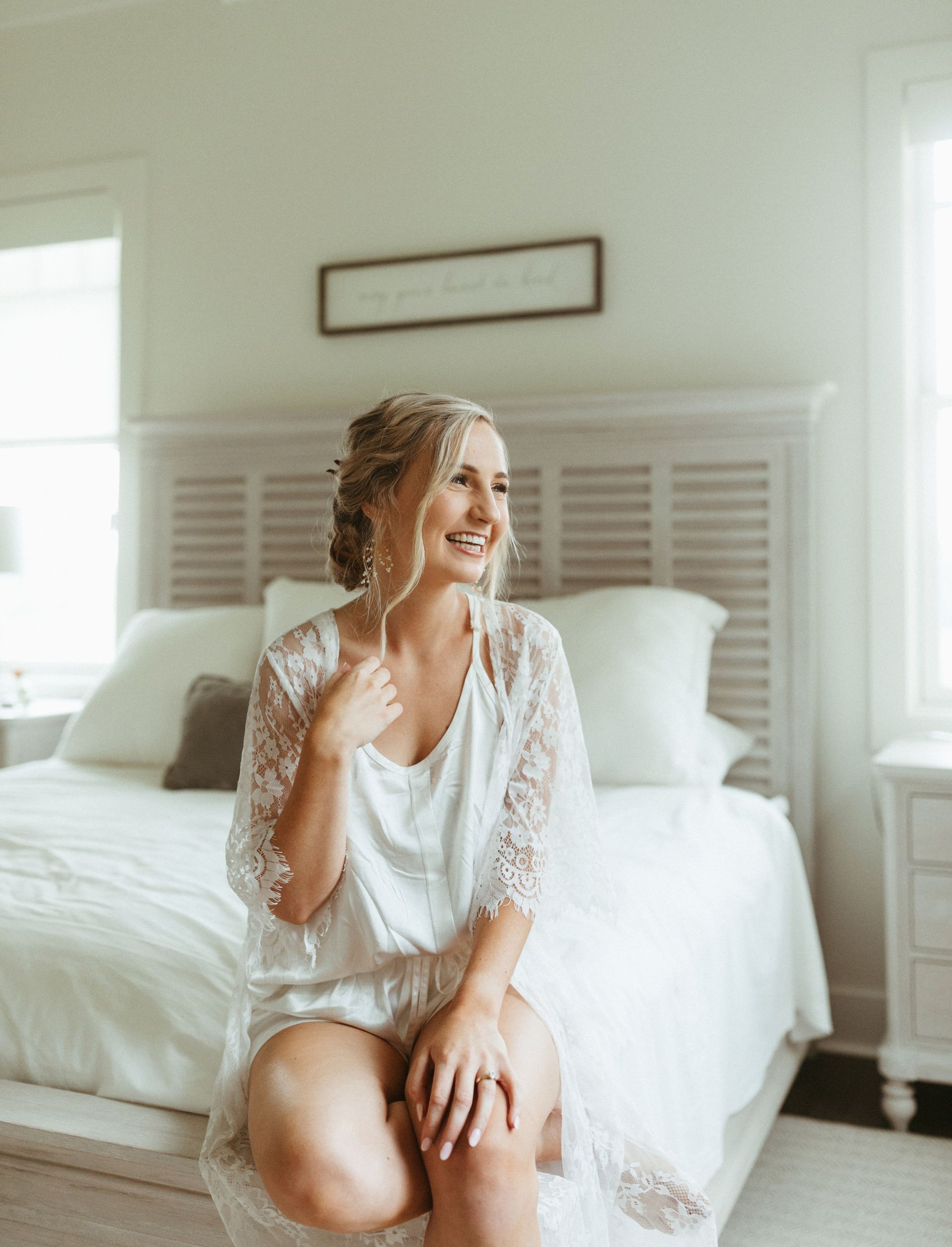 Woman in white lace outfit sits on a bed, smiling. Bedroom with light colors.