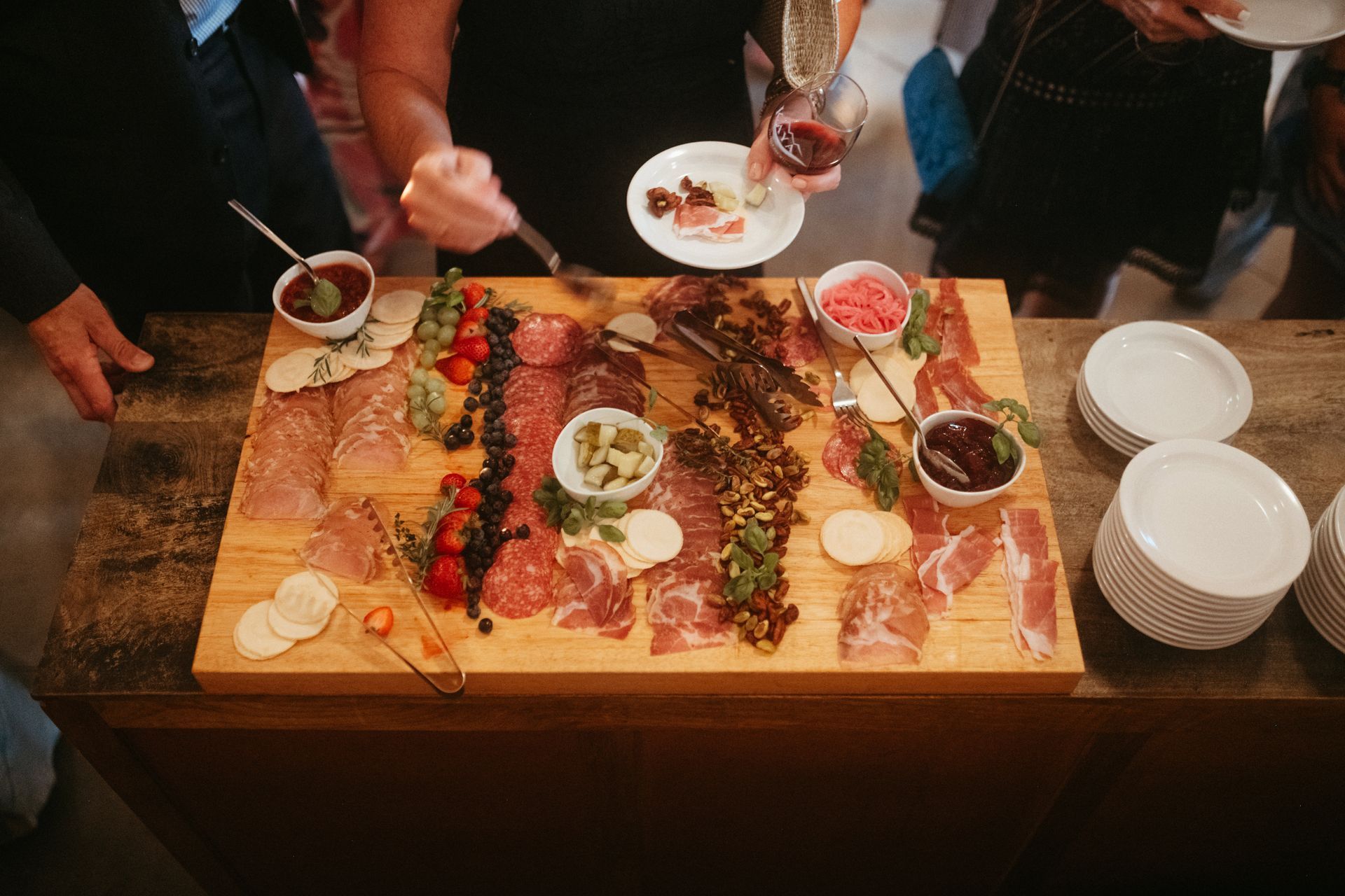 Large charcuterie board with assorted meats, cheeses, and garnishes, surrounded by people reaching for food.