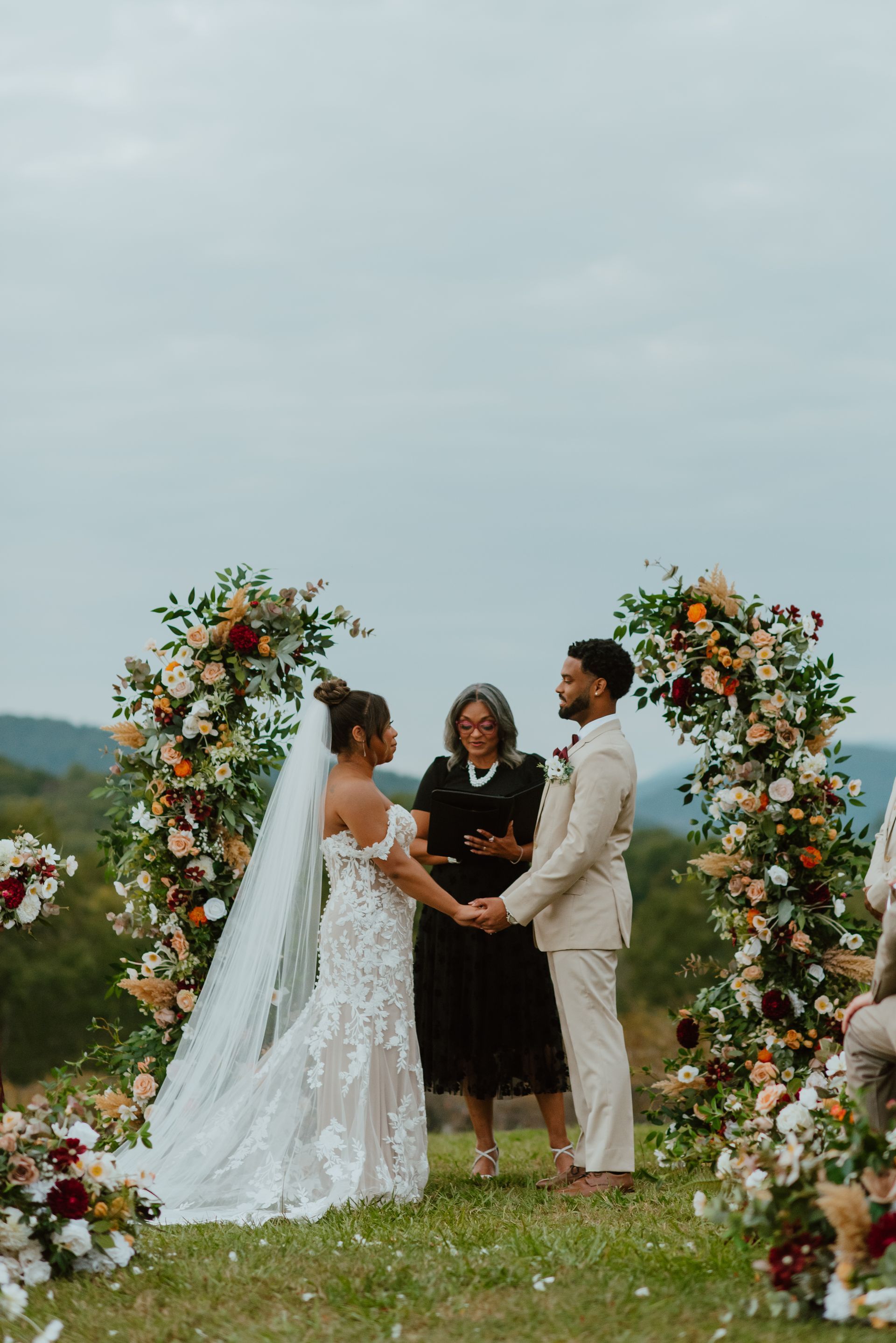 Couple holds hands at outdoor wedding ceremony; floral arch, officiant.
