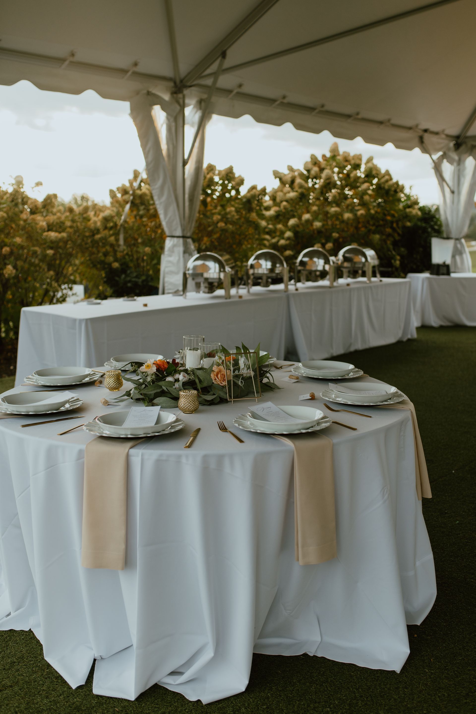 Round table set for a meal under a white tent, with a buffet table in the background.