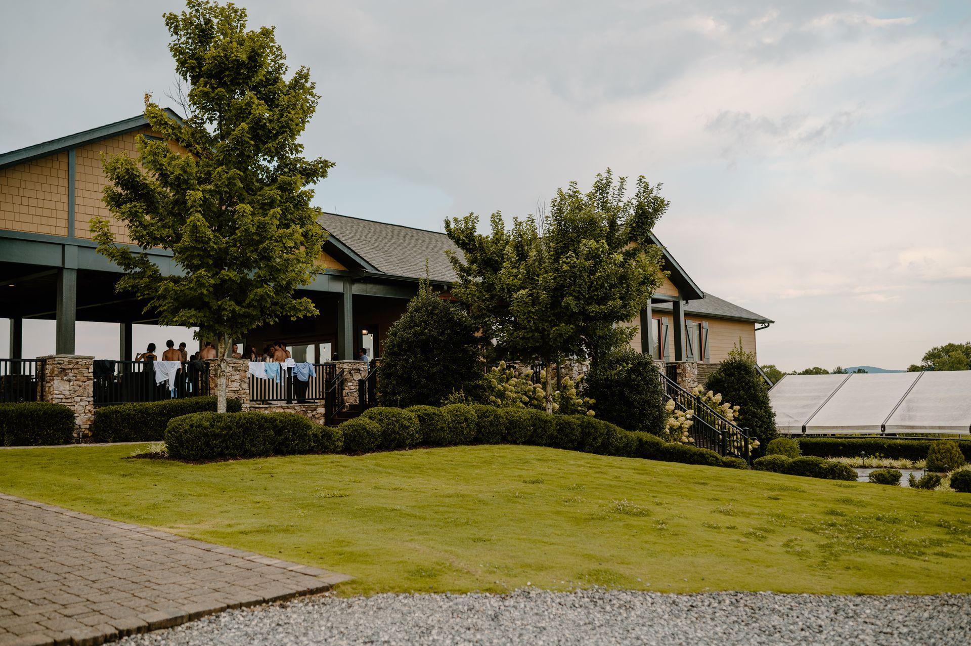 Exterior view of a building with a green lawn, trees, and a tent in the background under a cloudy sky.
