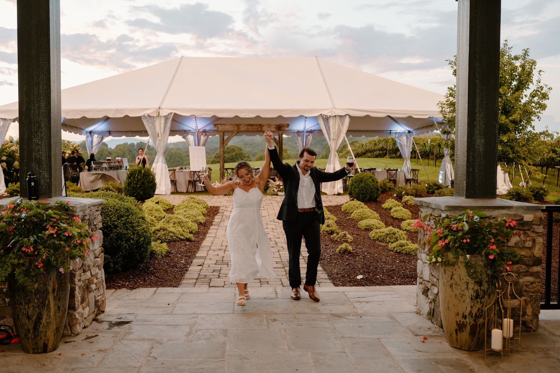 Newlyweds raising hands, joyfully exiting a stone walkway towards a tented reception, outdoor venue at sunset.