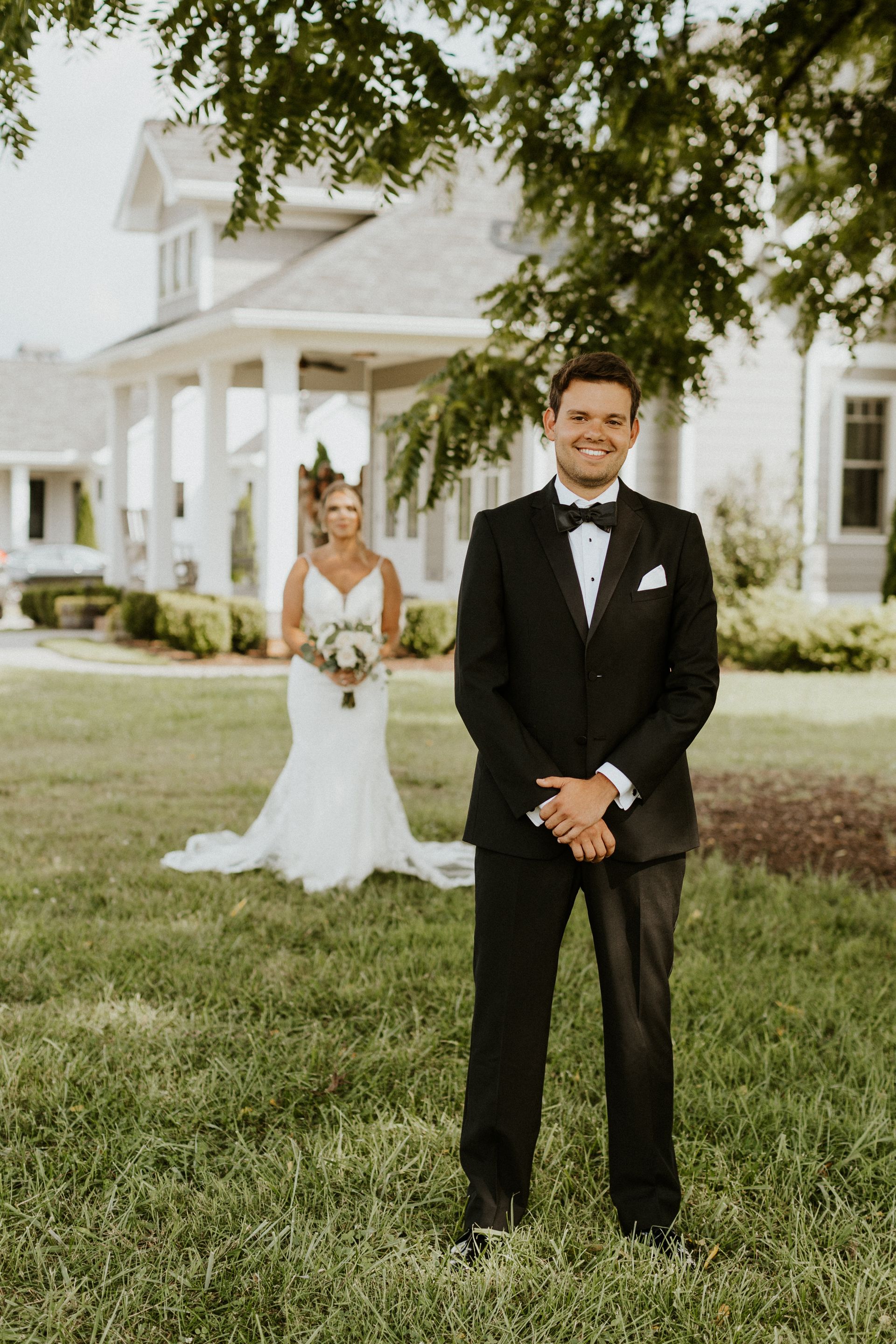Groom smiles, facing camera. Bride in white dress, holding flowers, stands behind him near house.