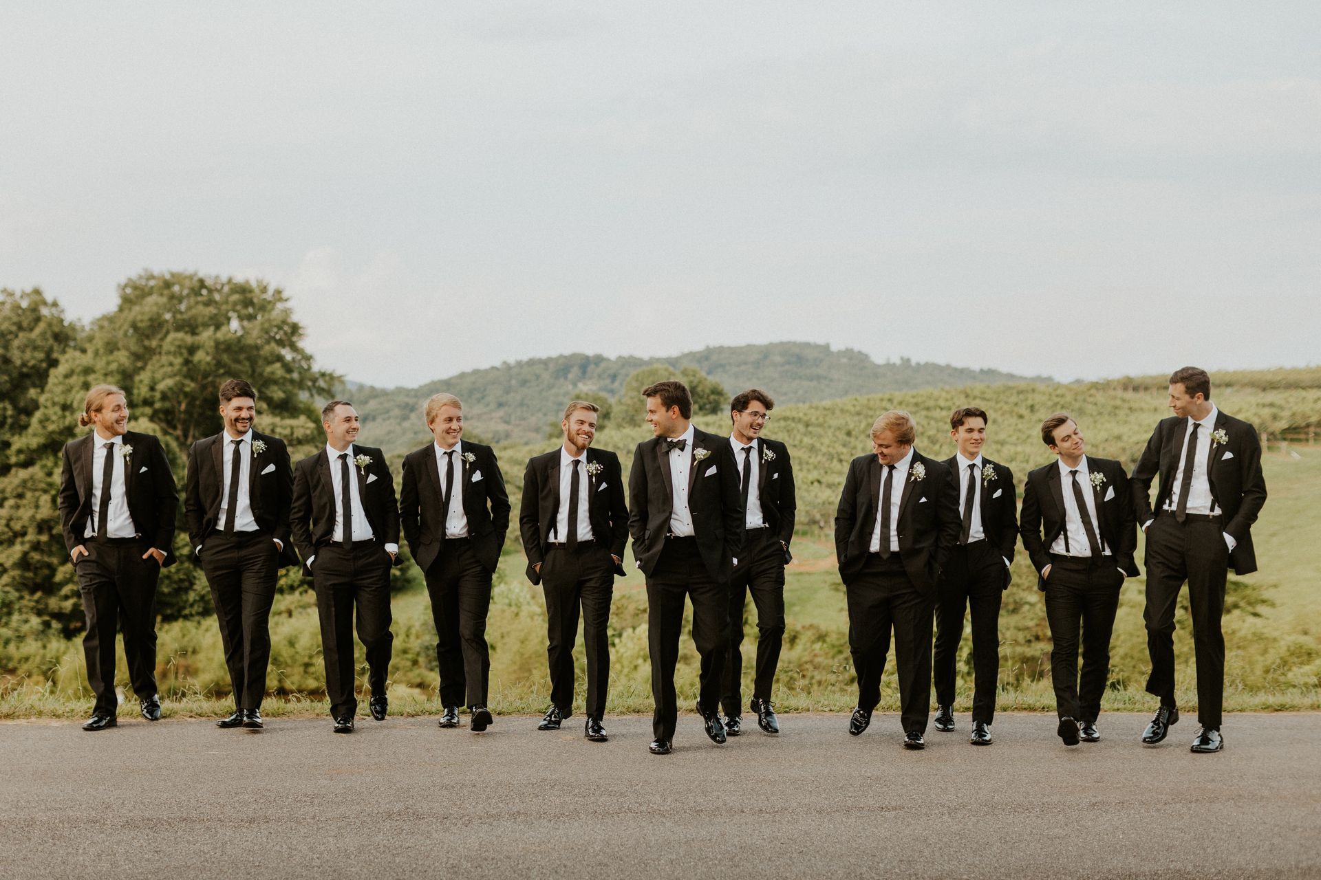 Group of men in black tuxedos walking on a road, laughing, with a green landscape in the background.