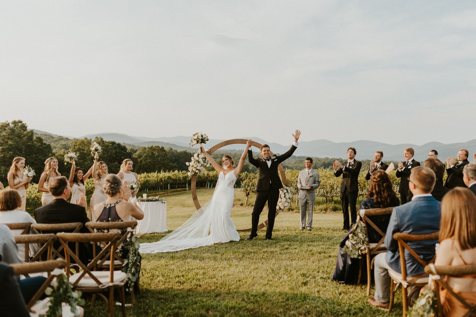 Newly married couple celebrates, arms raised, in front of a wedding arch, overlooking a vineyard. Guests applaud.