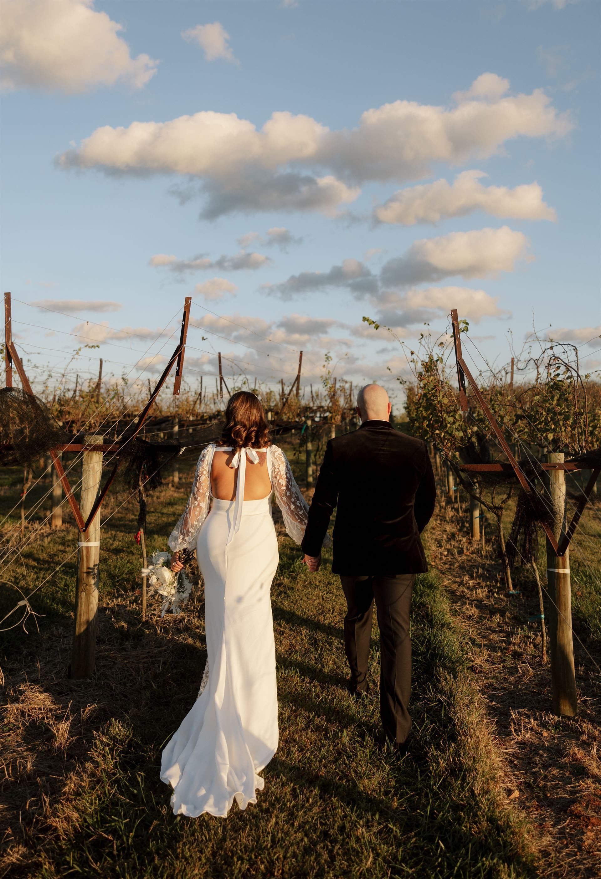 Bride and groom walking hand-in-hand through a vineyard at sunset.
