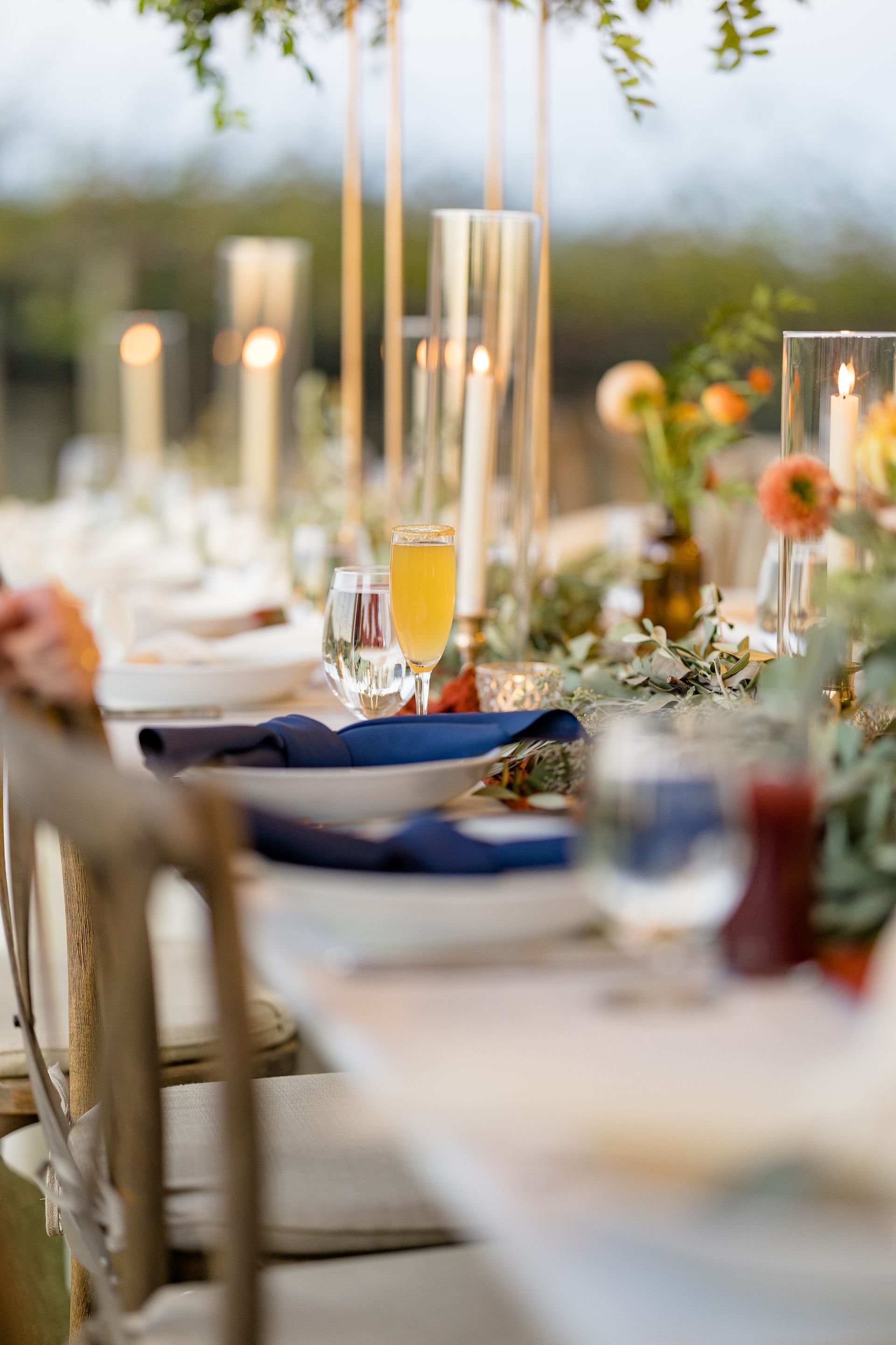 Elegant table setting with champagne flutes, navy napkins, and candles. Outdoors with blurred background.