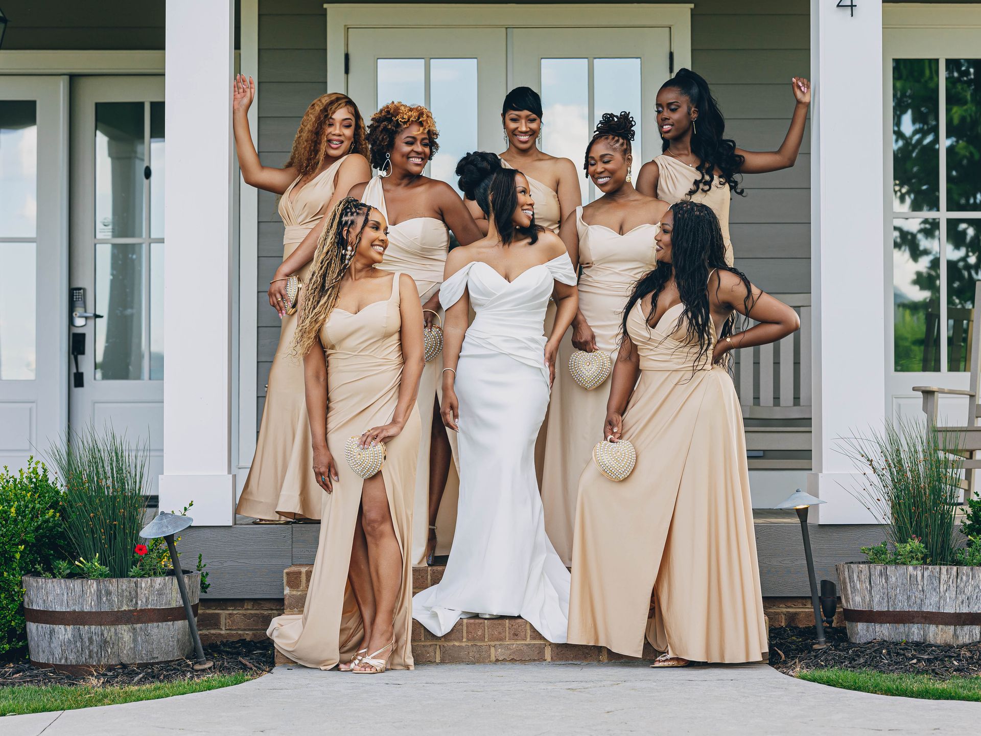 Bride in white gown poses with bridesmaids in tan dresses on porch.