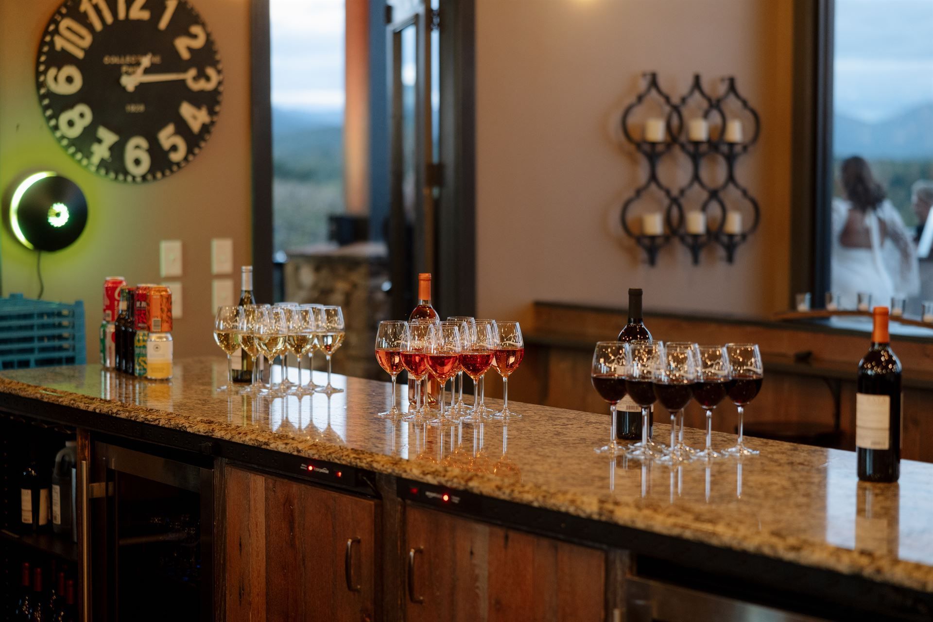 Bar with wine glasses and bottles on counter, large clock, decorative wall sconces, view of outdoor setting.