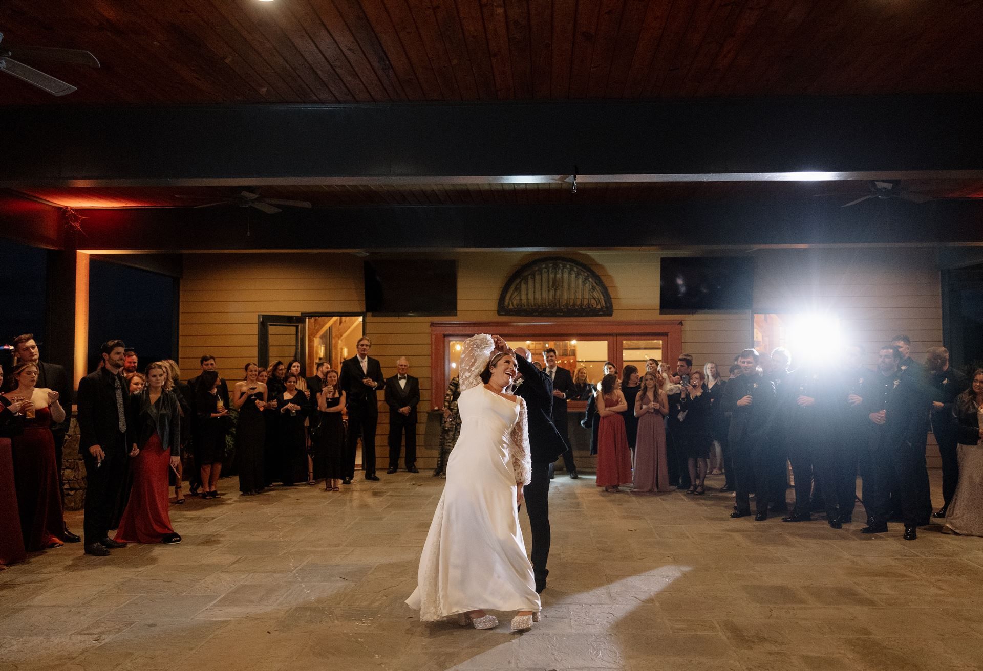 Couple dancing at wedding reception. Guests watch, lit by outdoor lights.