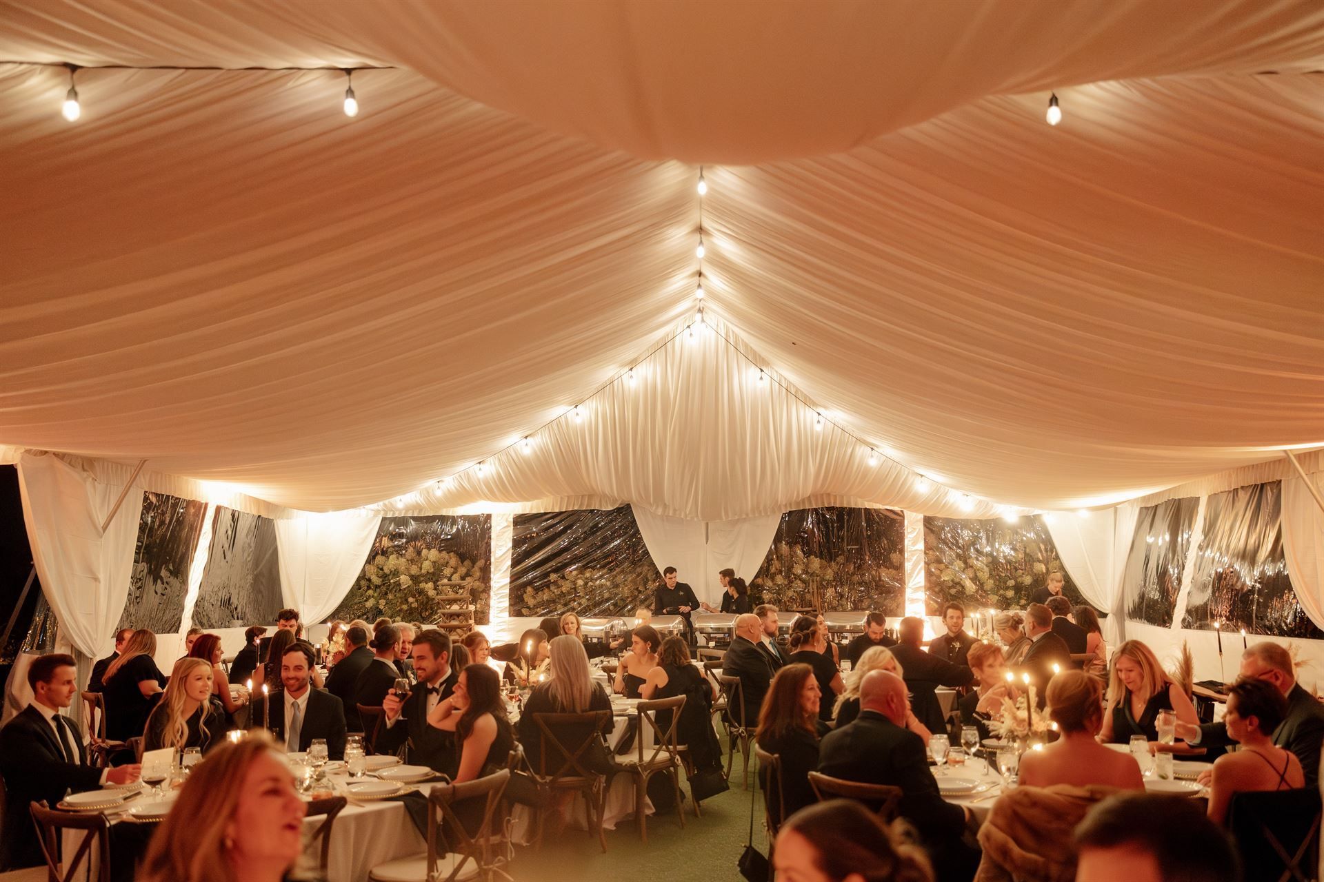People seated at tables in a tent, lit with string lights. Evening event.