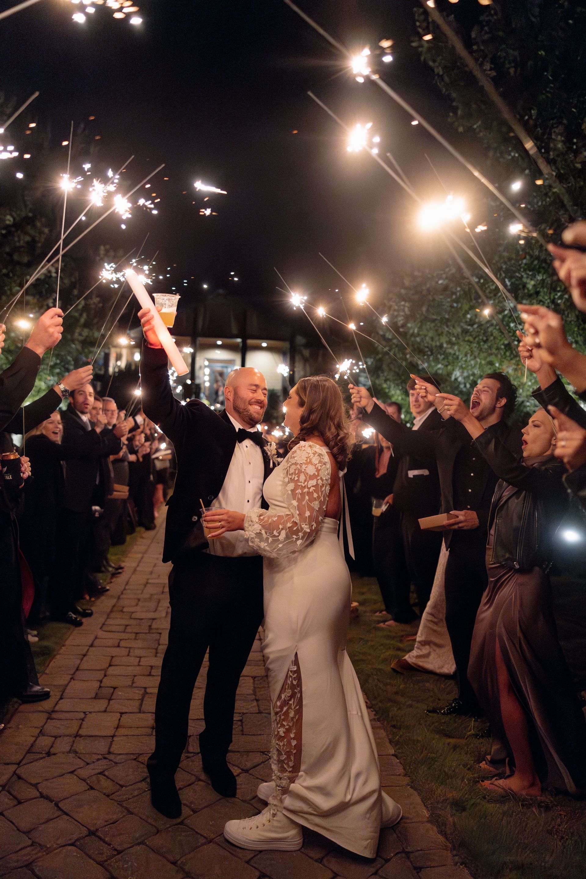Newlyweds amid a sparkler send-off, embracing and smiling at night, framed by onlookers and a brick path.