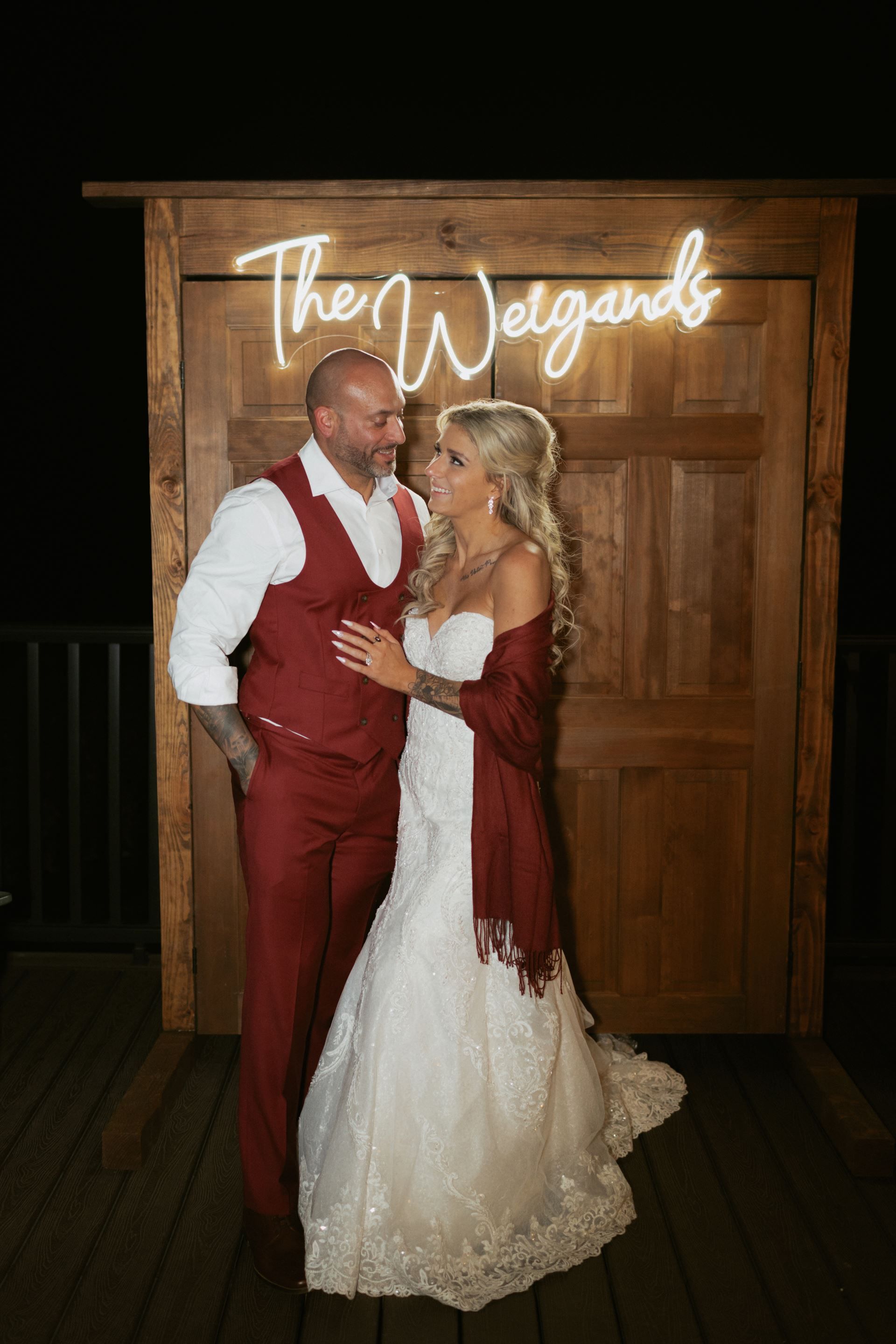 Newlyweds pose in front of a wooden backdrop with a neon sign that says 