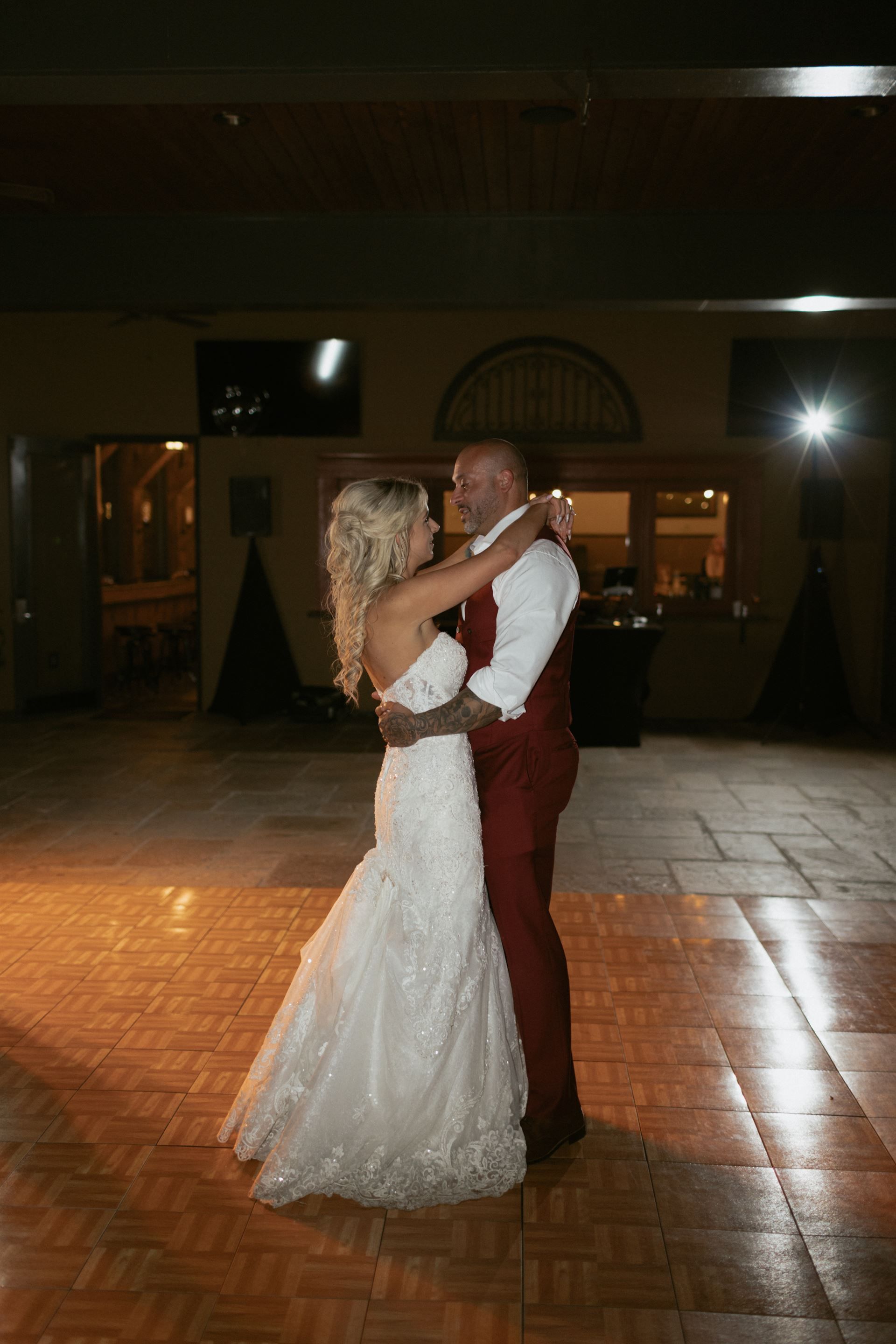 Bride and groom dance on a wooden floor at their wedding reception, embracing and smiling.