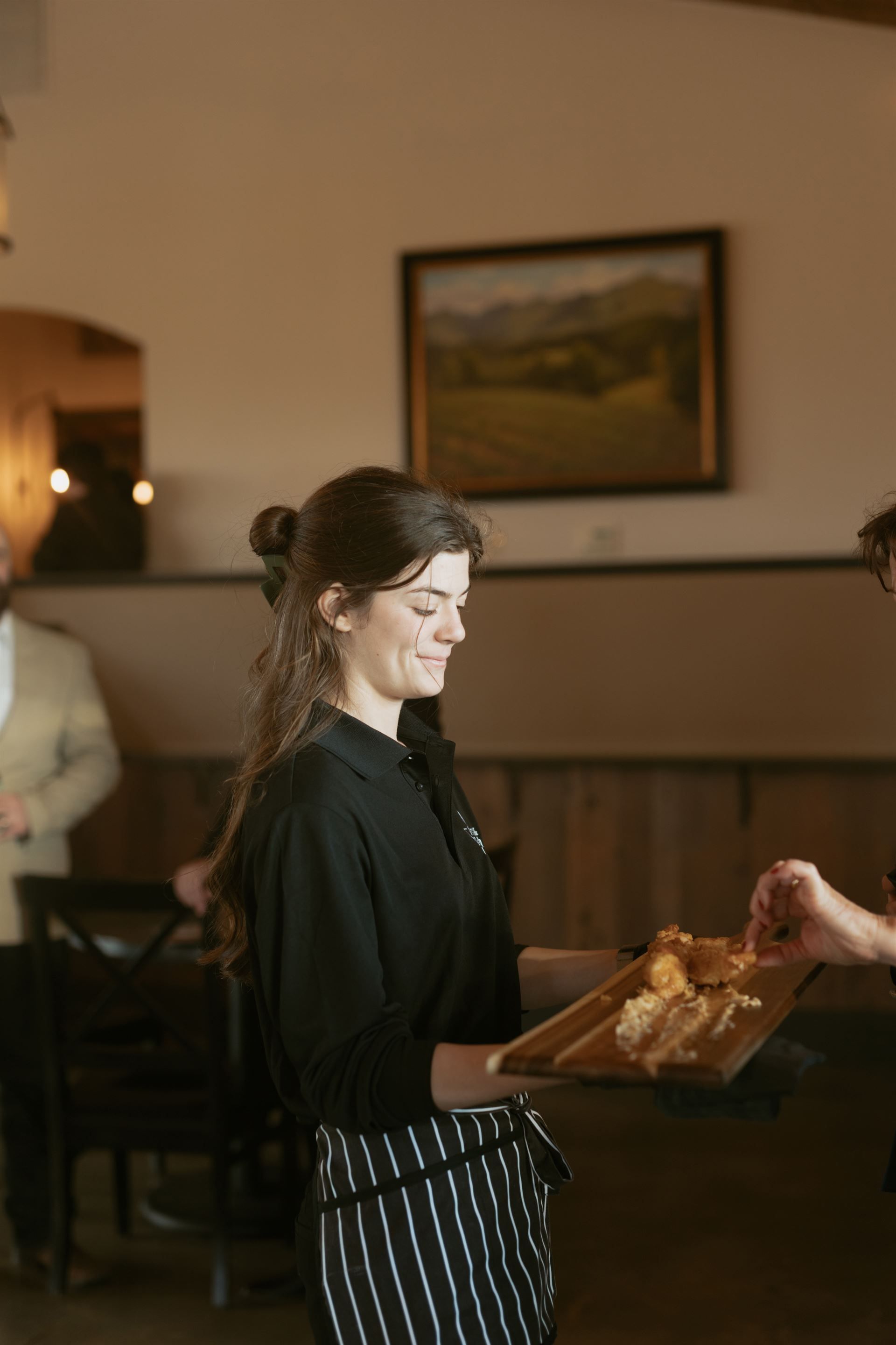 Waitress carrying a wooden tray with food inside a restaurant; she smiles while another person reaches for a food item.