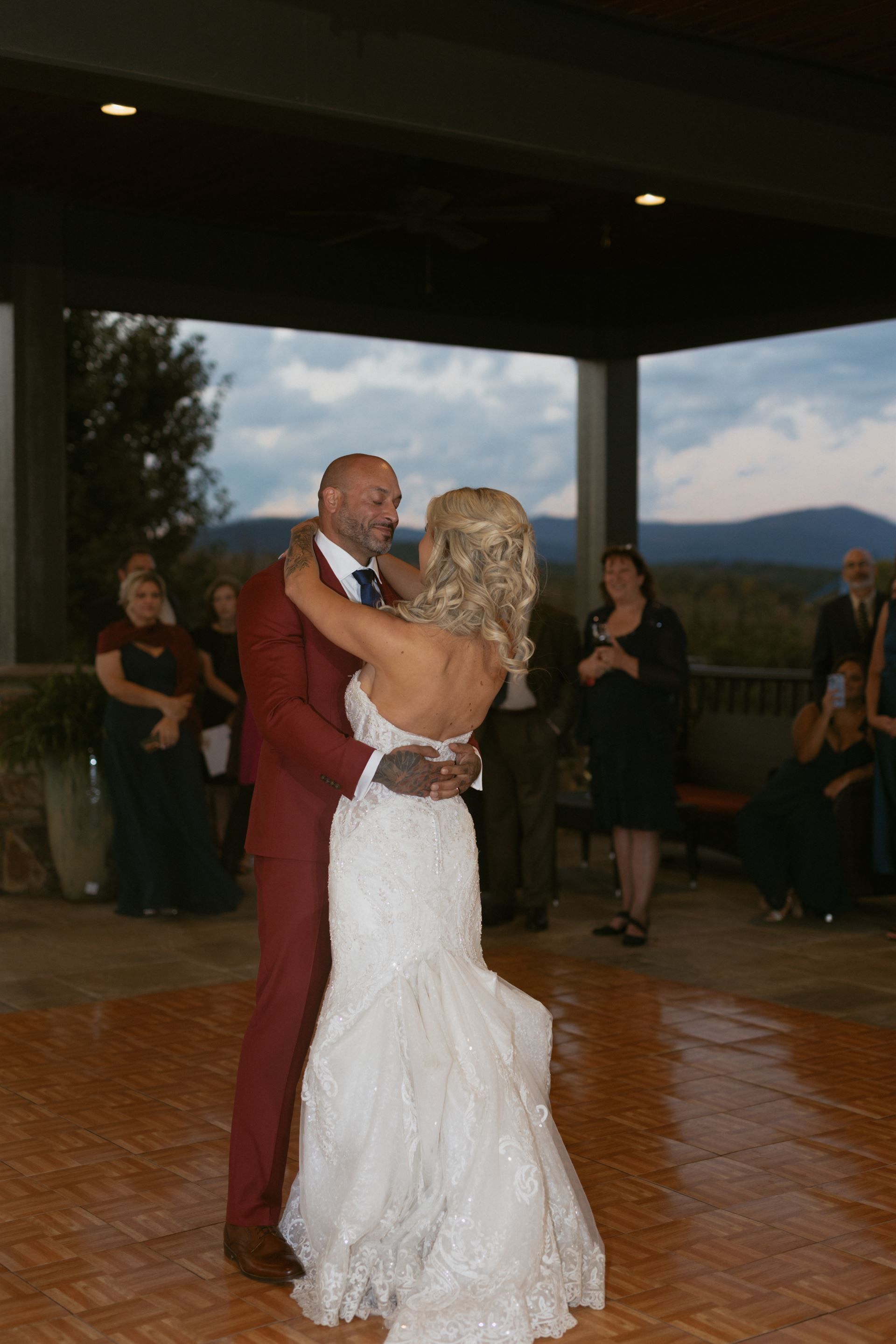 Bride and groom dancing at a wedding reception, she in a white gown, he in a red suit, outdoors under a covered space.