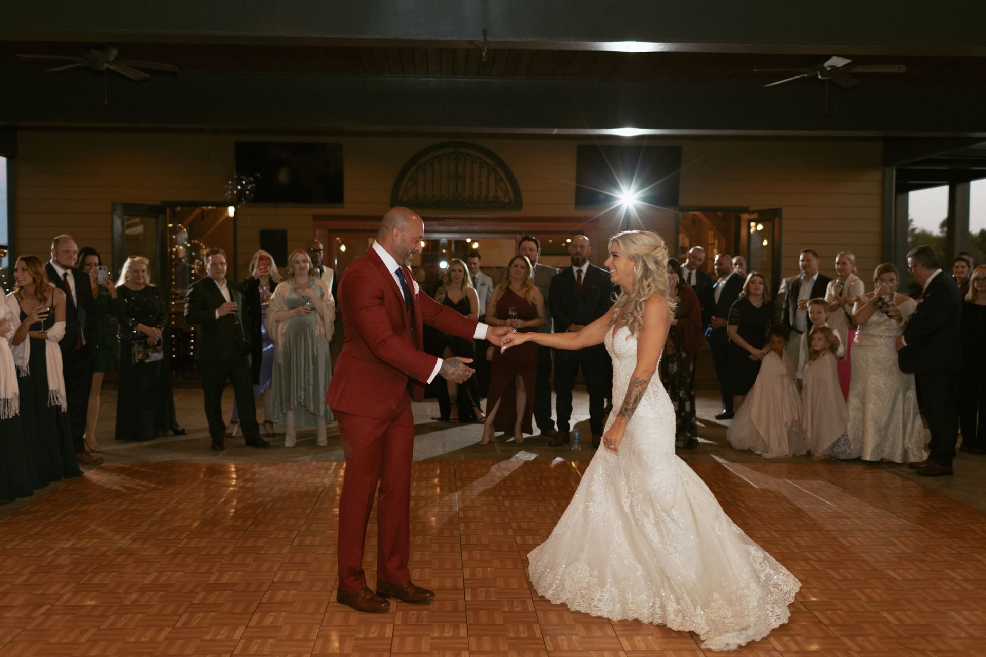 Bride and groom dance at reception; onlookers watch. Groom in red suit; bride in white gown. Wooden dance floor.